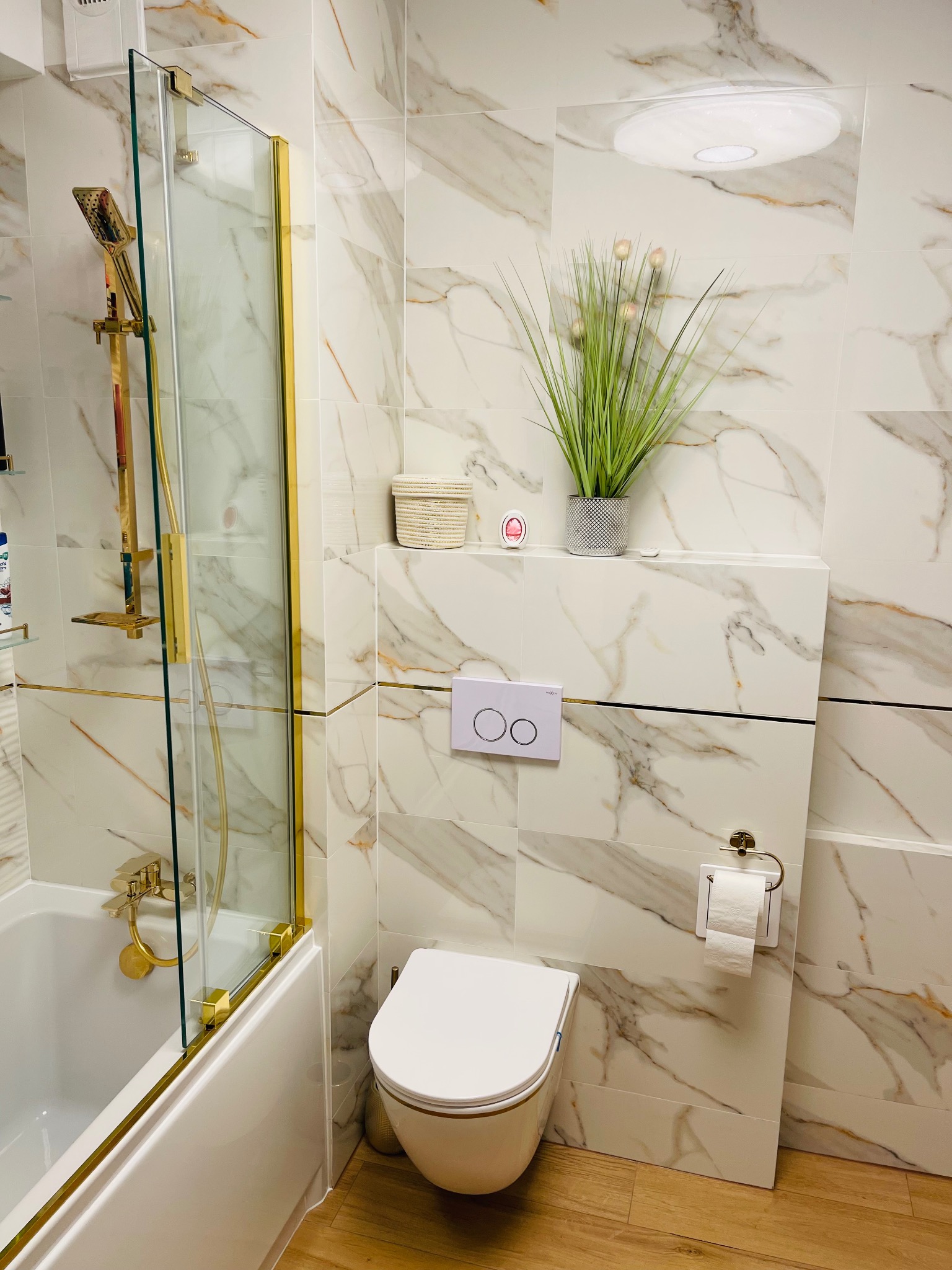 Elegancka łazienka z marmurowymi płytkami, złotymi akcentami na kabinie prysznicowej i armaturze, wisząca toaleta z białym przyciskiem spłukującym i dekoracyjna roślina w doniczce.