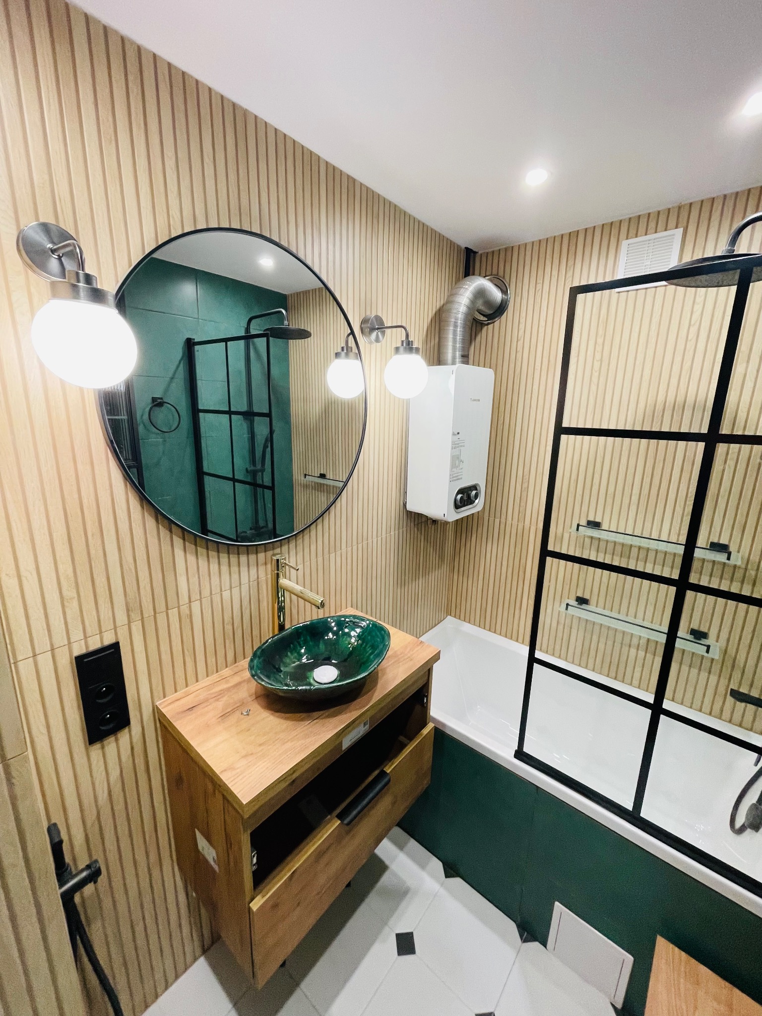 Nowoczesna łazienka z drewnianymi panelami ściennymi, okrągłym lustrem z podświetleniem, zieloną umywalką nablatową i kabiną prysznicową typu walk-in z czarnymi profilami, widoczny bojler.