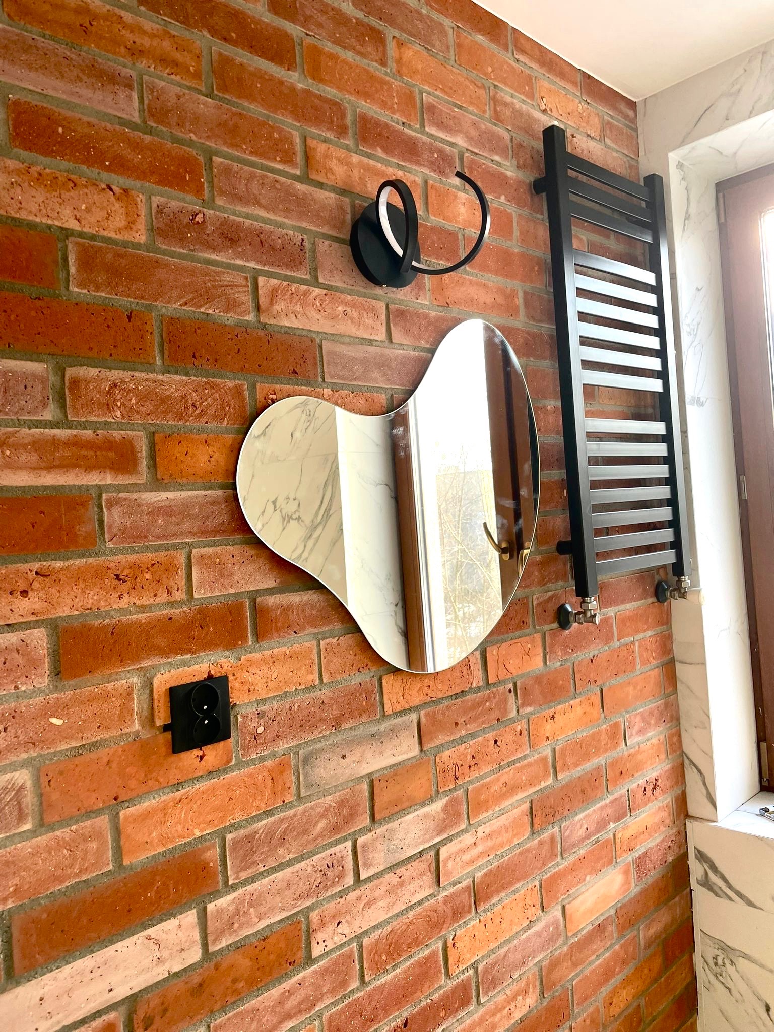 Ceglana ściana w łazience z asymetrycznym lustrem, czarnym grzejnikiem drabinkowym i kinkietem o abstrakcyjnym kształcie.