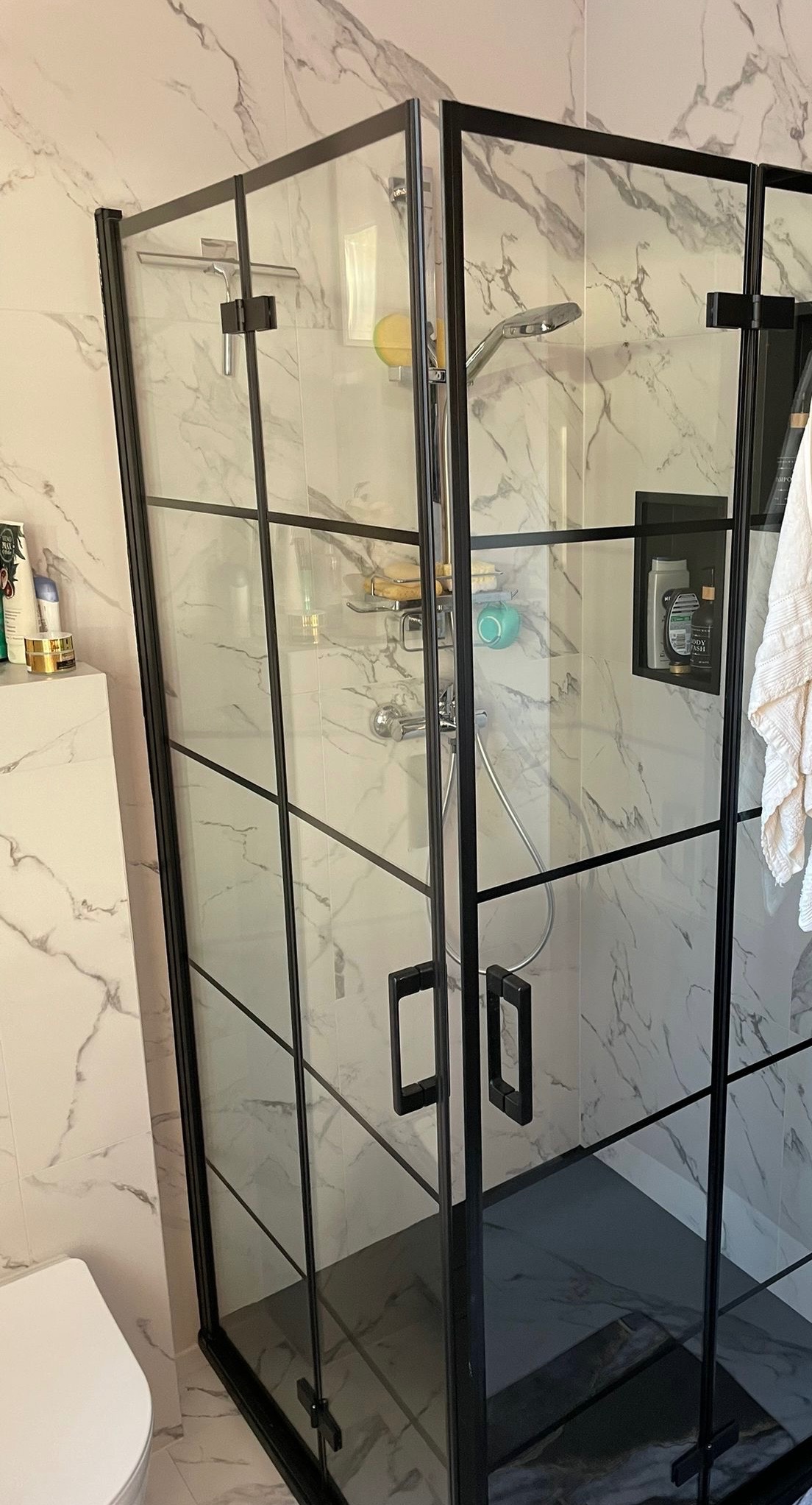 Nowoczesna kabina prysznicowa z czarnymi profilami na tle marmurowych płytek, widoczna bateria, półka na kosmetyki w ścianie i akcesoria prysznicowe.