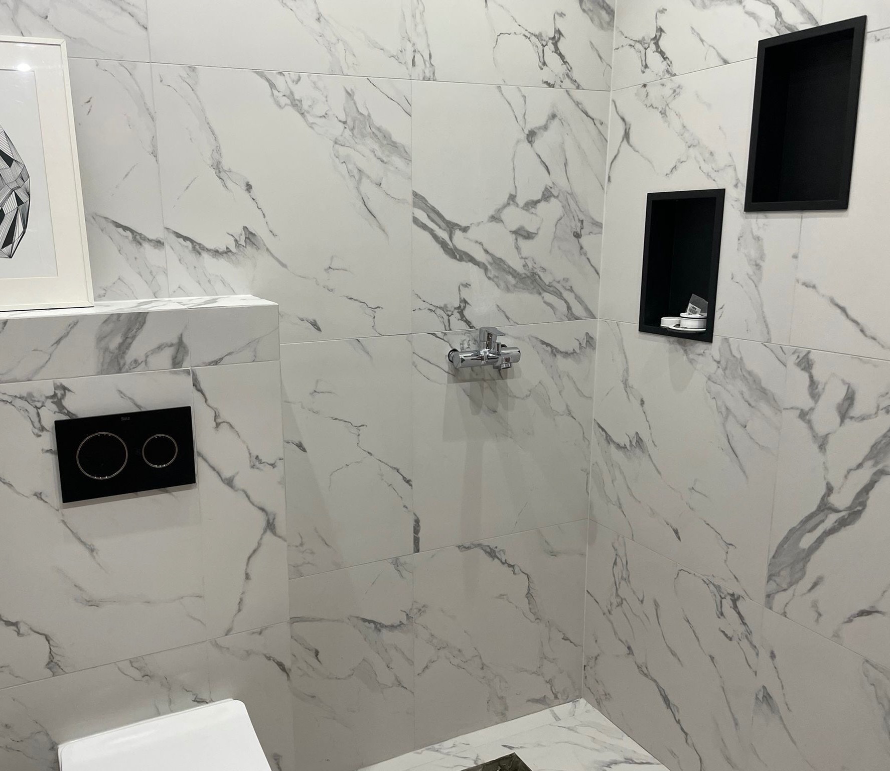 Białe marmurowe płytki w łazience z czarnymi wnękami i panelem przycisku spłukiwania, minimalistyczny design, armatura chromowana.