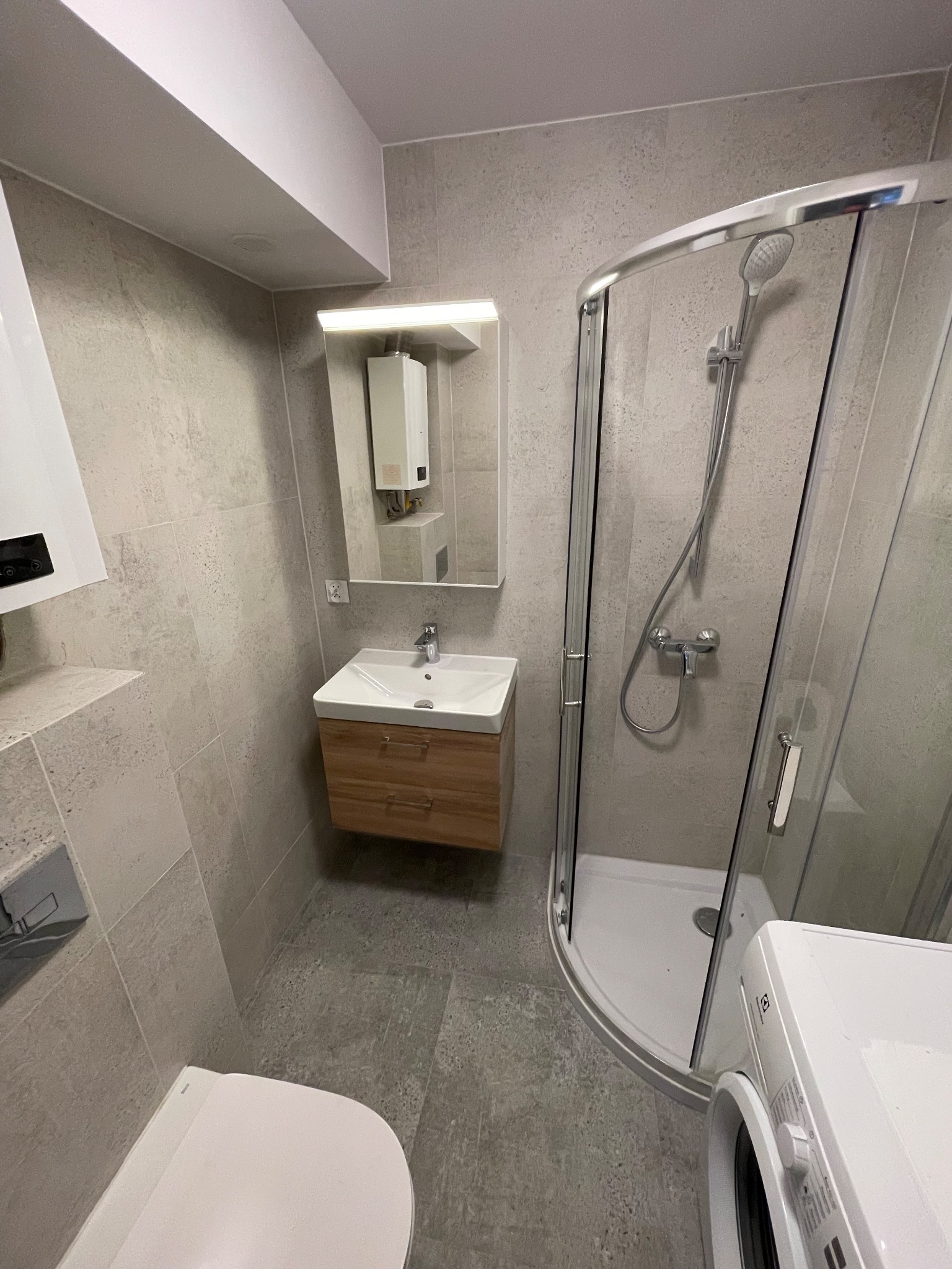 Nowoczesna łazienka w szarościach z kabiną prysznicową z zaokrąglonymi drzwiami, umywalką z szafką z jasnego drewna, podwieszanym WC i pralką Electrolux.