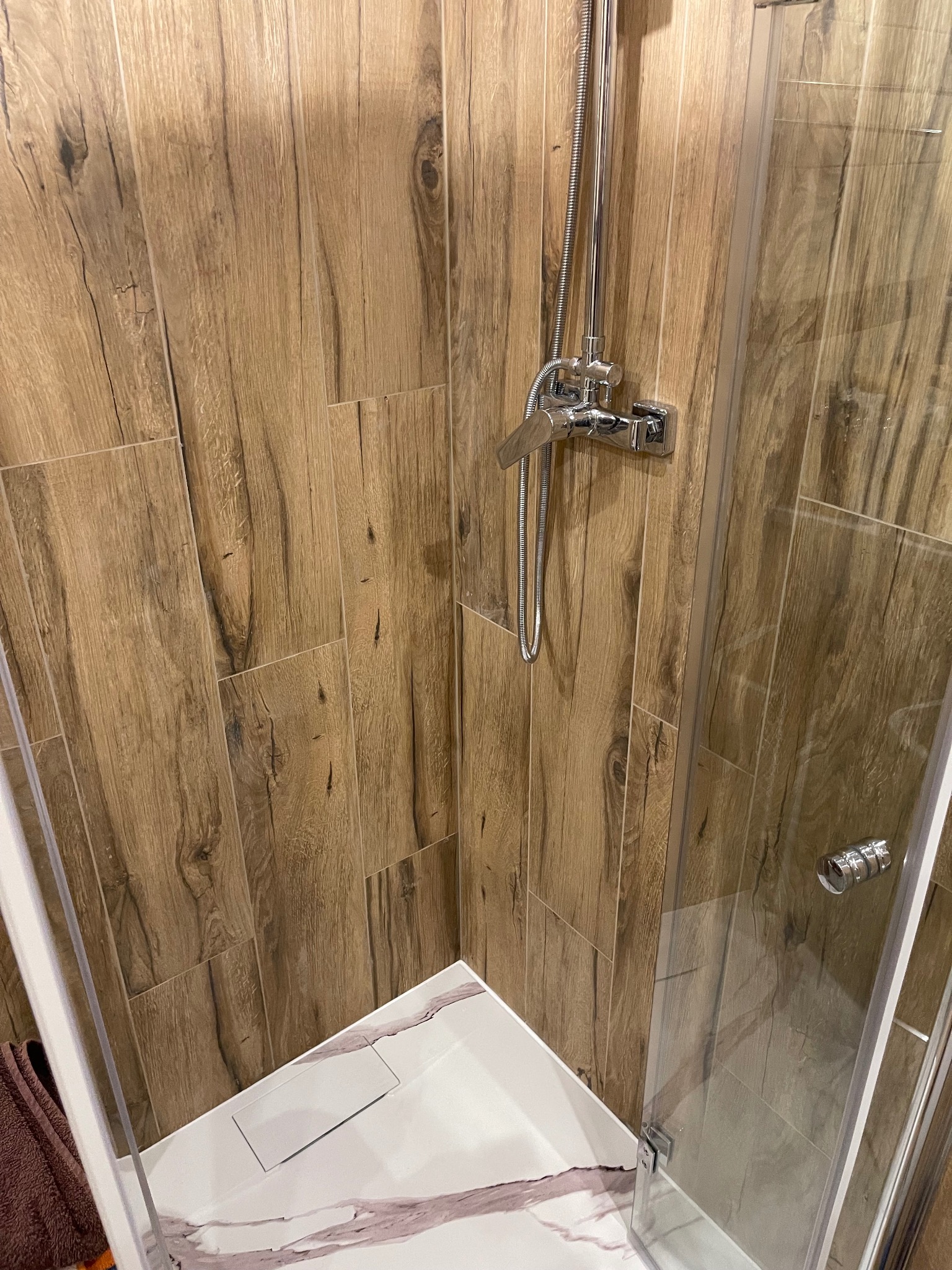 Nowoczesna kabina prysznicowa z armaturą chromowaną i imitującymi drewno płytkami ściennymi. Biały brodzik z szarymi żyłkami i odpływem liniowym.