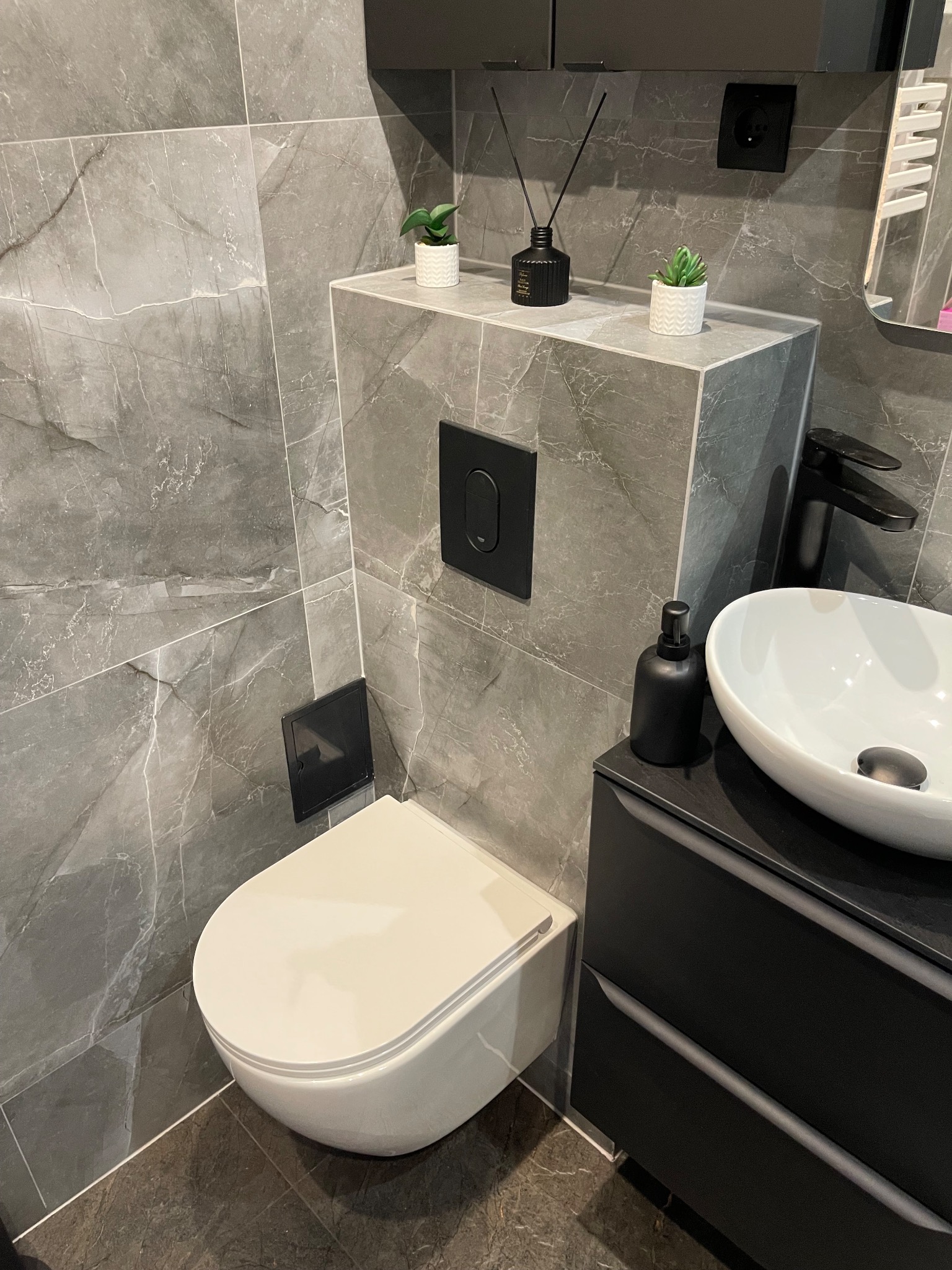 Nowoczesna łazienka z marmurowymi płytkami, podwieszaną toaletą i umywalką nablatową. Czarna armatura i dodatki, minimalistyczny design. Drobne rośliny doniczkowe jako akcent dekoracyjny.