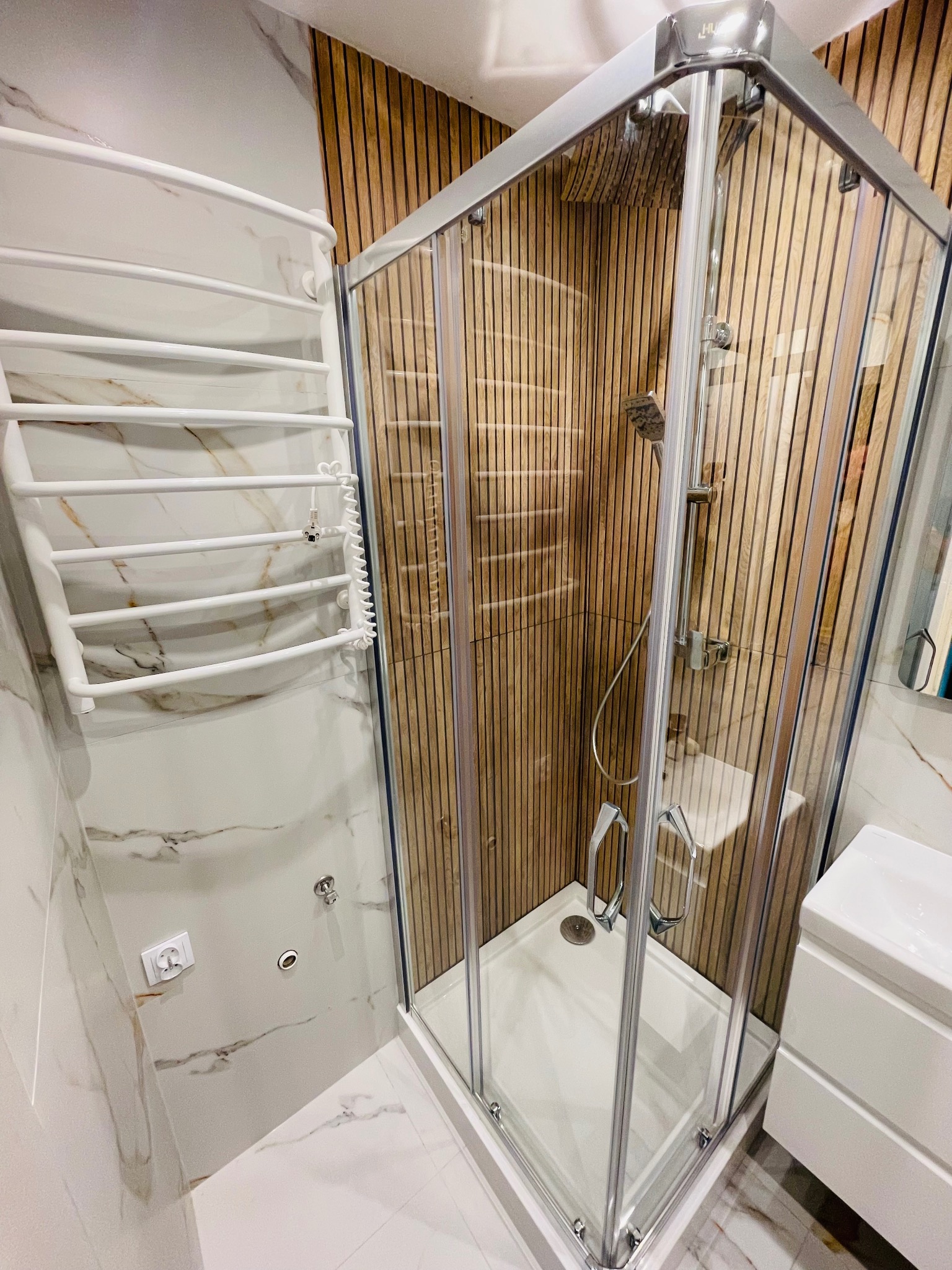 Nowoczesna łazienka z kabiną prysznicową w rogu, wykończona drewnianą okładziną. Białe marmurowe płytki i grzejnik drabinkowy. Widok na umywalkę i białą podłogę.