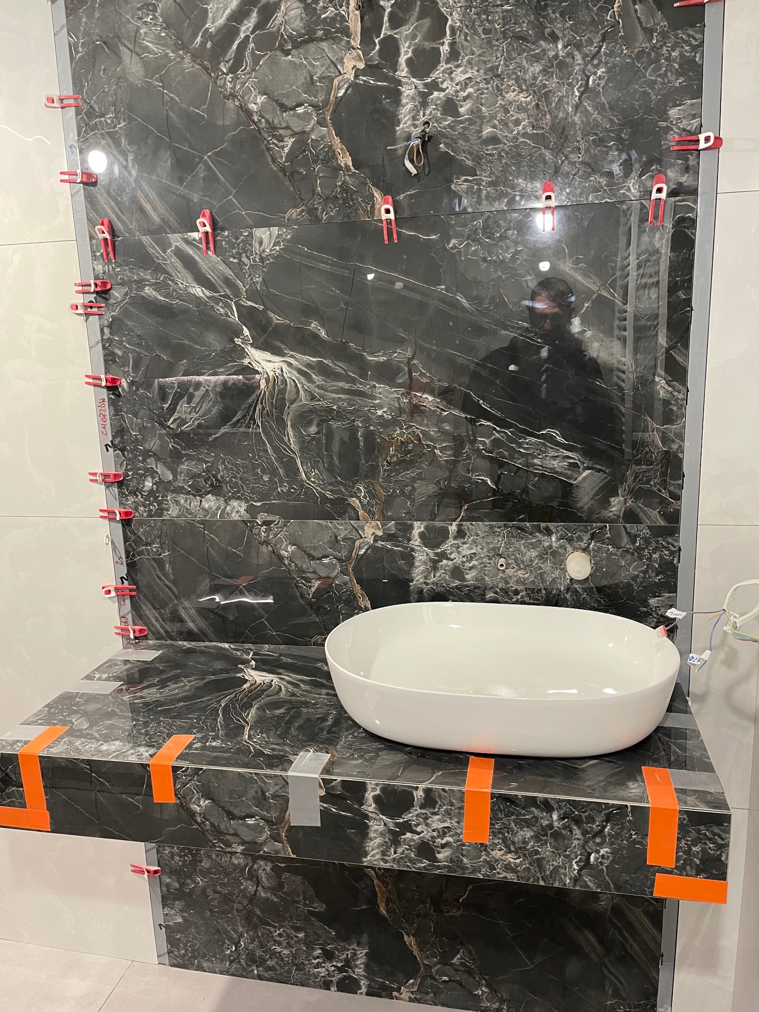 Montaż czarnych płytek imitujących marmur w łazience, z użyciem klipsów poziomujących. Widoczna biała umywalka nablatowa i odbicie osoby w płytkach.