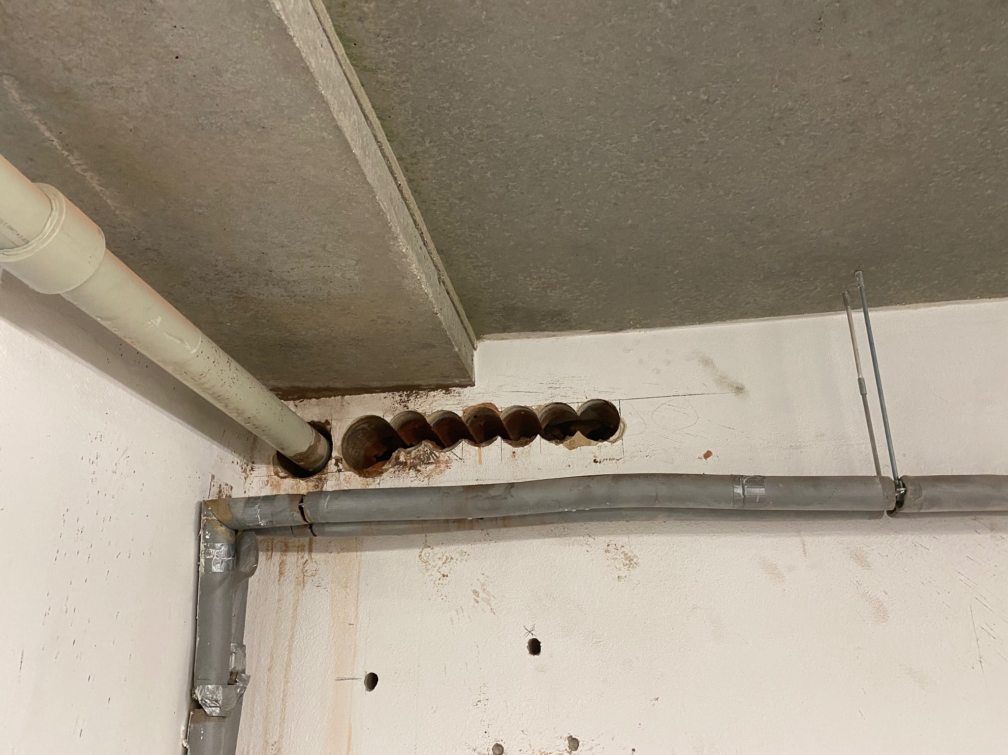 Betonowy sufit z rzędem wywierconych otworów, obok widoczne rury instalacyjne, ściany z widocznymi śladami wiercenia.