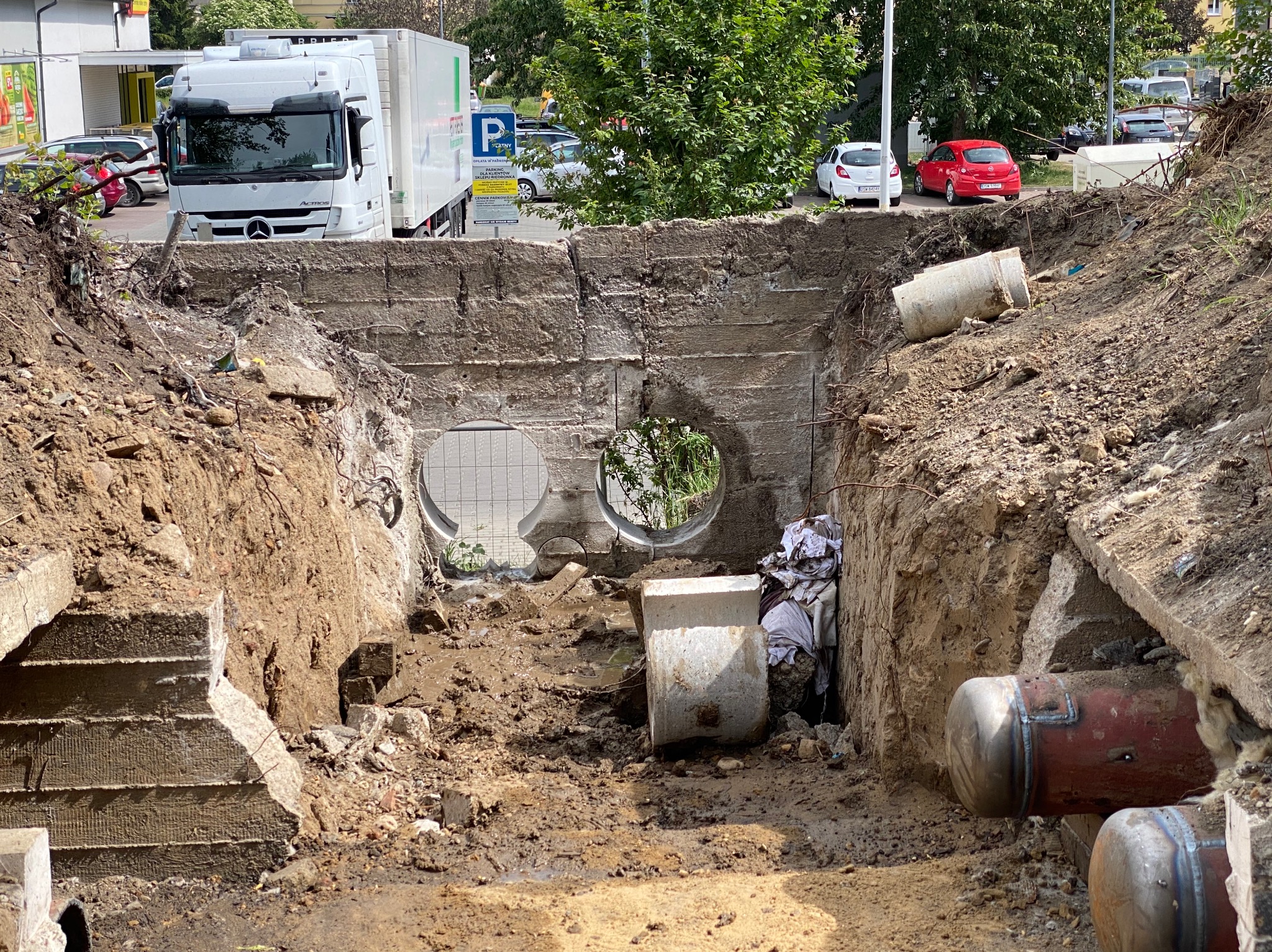 Fragment wykopu z widoczną ścianą betonową z dwoma okrągłymi otworami, na górze widoczny biały samochód ciężarowy, w tle zaparkowane samochody i drzewa.