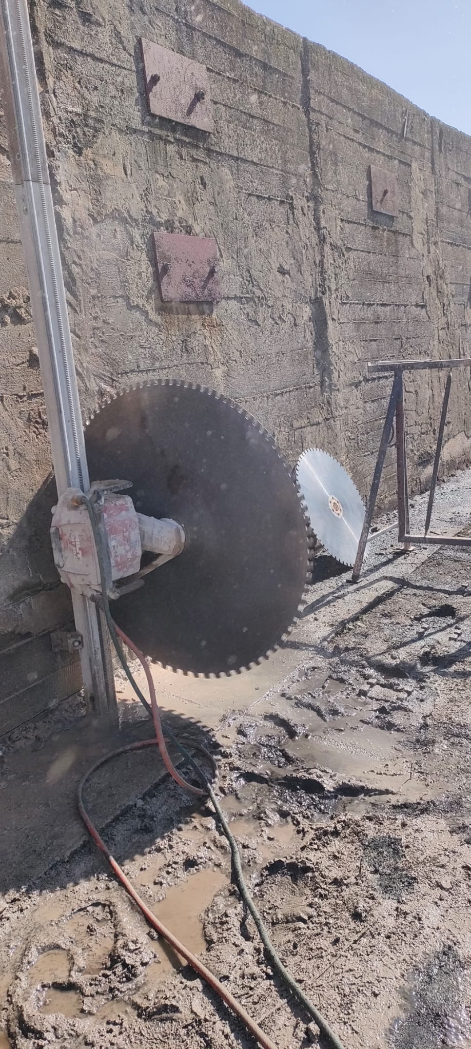 Piła diamentowa do betonu przymocowana do szyny, tnąca grubą ścianę betonową, w tle druga piła i metalowa konstrukcja, błoto na ziemi.