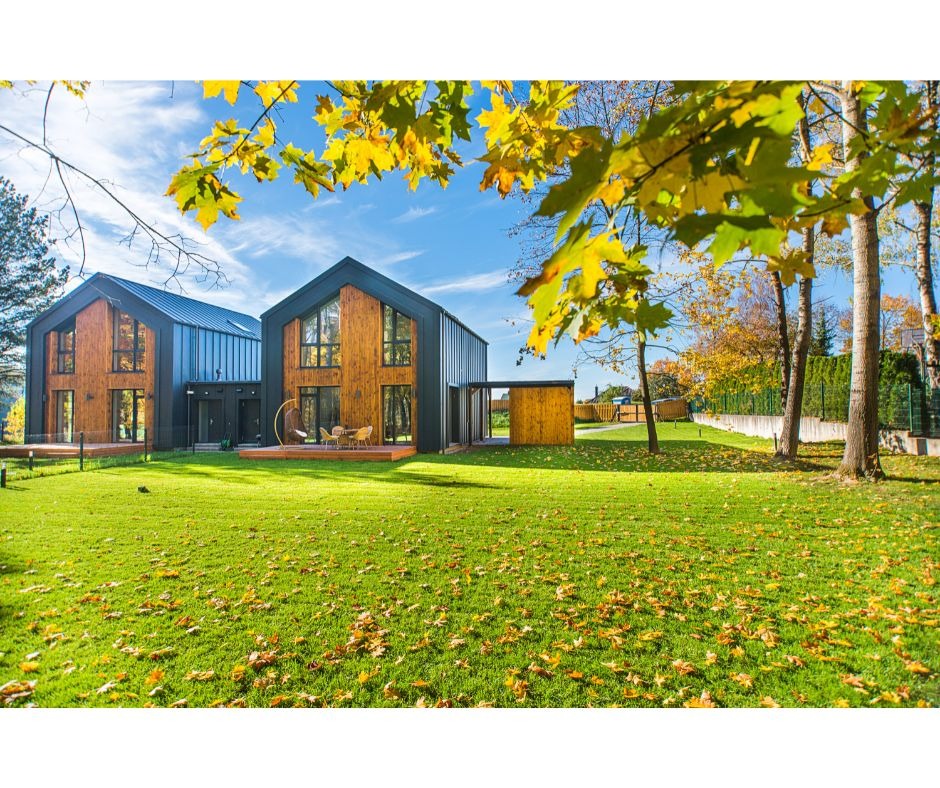 Dwa nowoczesne budynki z drewnianą elewacją i ciemnym dachem, połączone drewnianym pomostem, otoczone zielonym trawnikiem z opadłymi liśćmi jesiennymi, widok z perspektywy trawnika