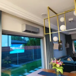 Nowoczesny klimatyzator ścienny w jasnym salonie z widokiem na ogród przez duże okno. Wnętrze dopełnia designerska lampa i eleganckie zasłony.