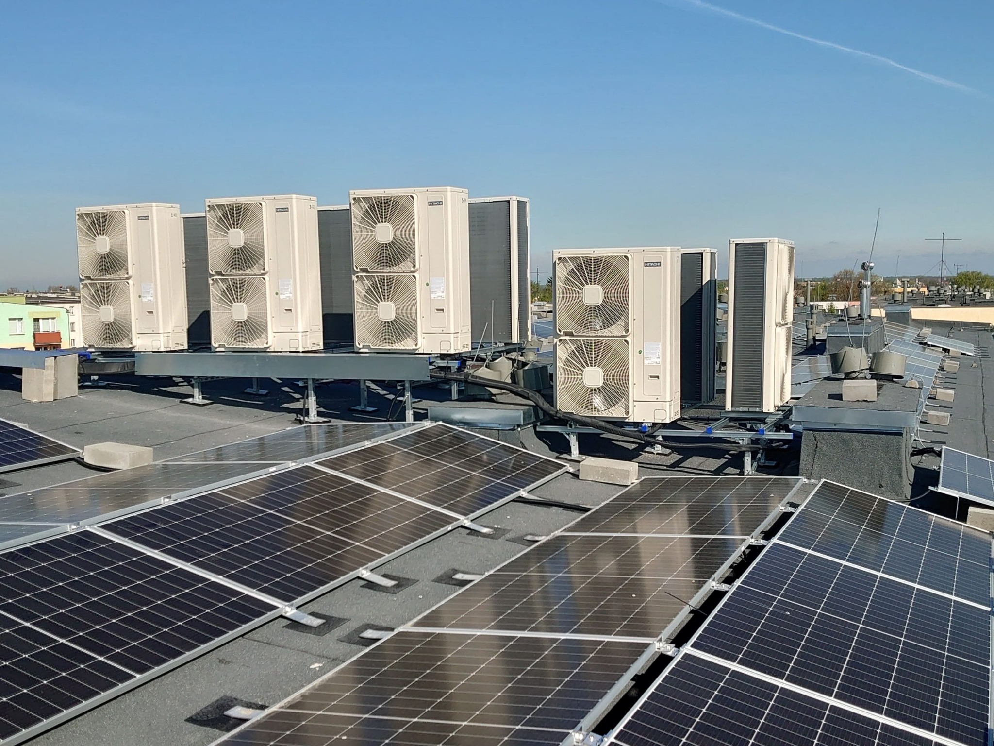 Instalacja klimatyzacji Hitachi składająca się z wielu jednostek zewnętrznych na dachu budynku, obok paneli fotowoltaicznych w słoneczny dzień.