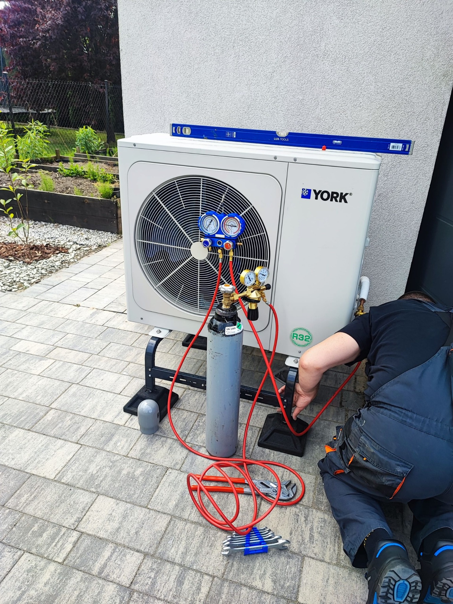 Serwisant w kombinezonie podłącza butlę z czynnikiem chłodniczym R32 do zewnętrznej jednostki pompy ciepła York, poziomica leży na obudowie, na ziemi leżą klucze.