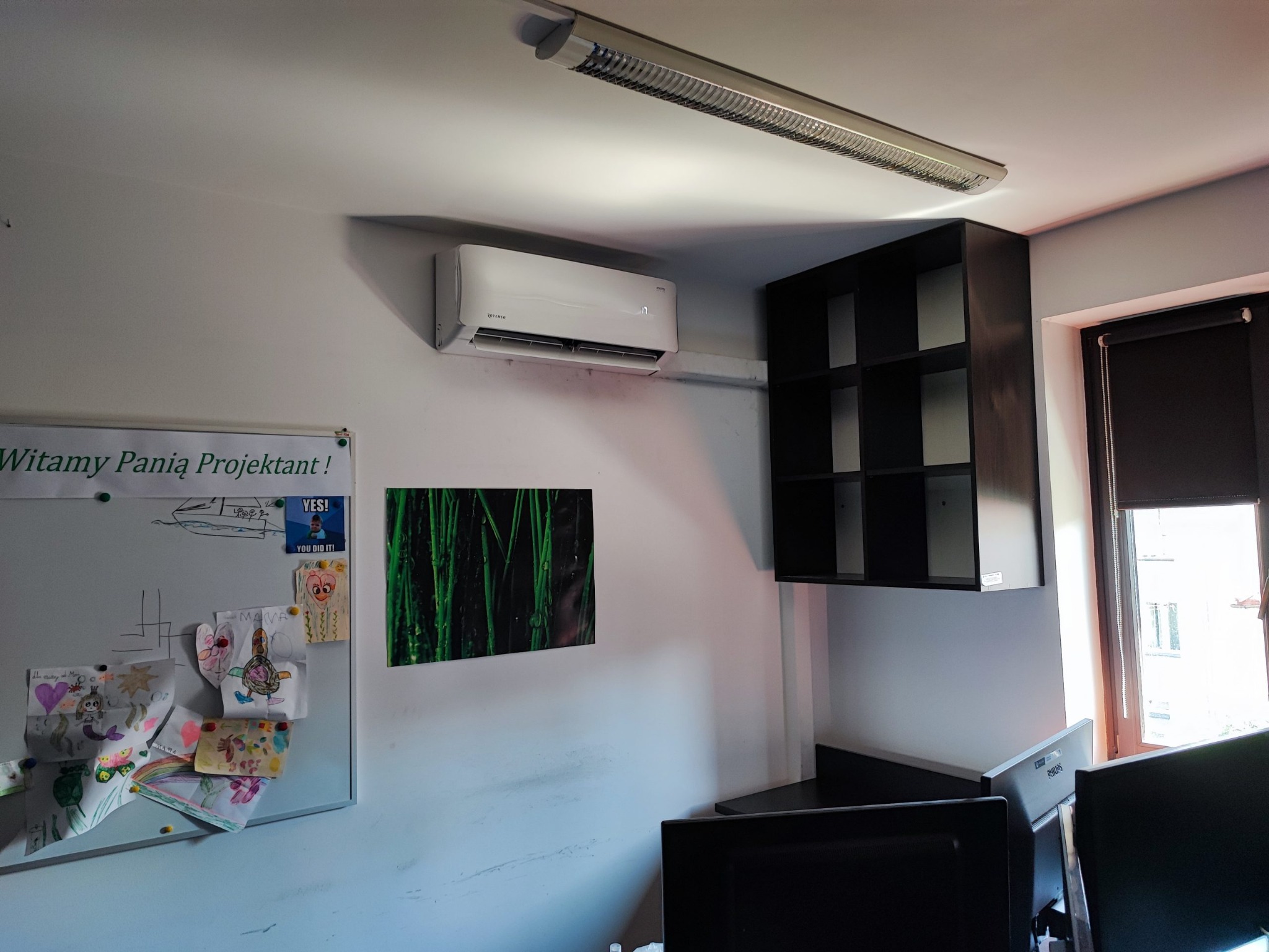 Klimatyzator Rotenso zamontowany w biurze, widoczny podłużny panel LED. Na ścianie tablica z rysunkami i napis 'Witamy Panią Projektant!'.