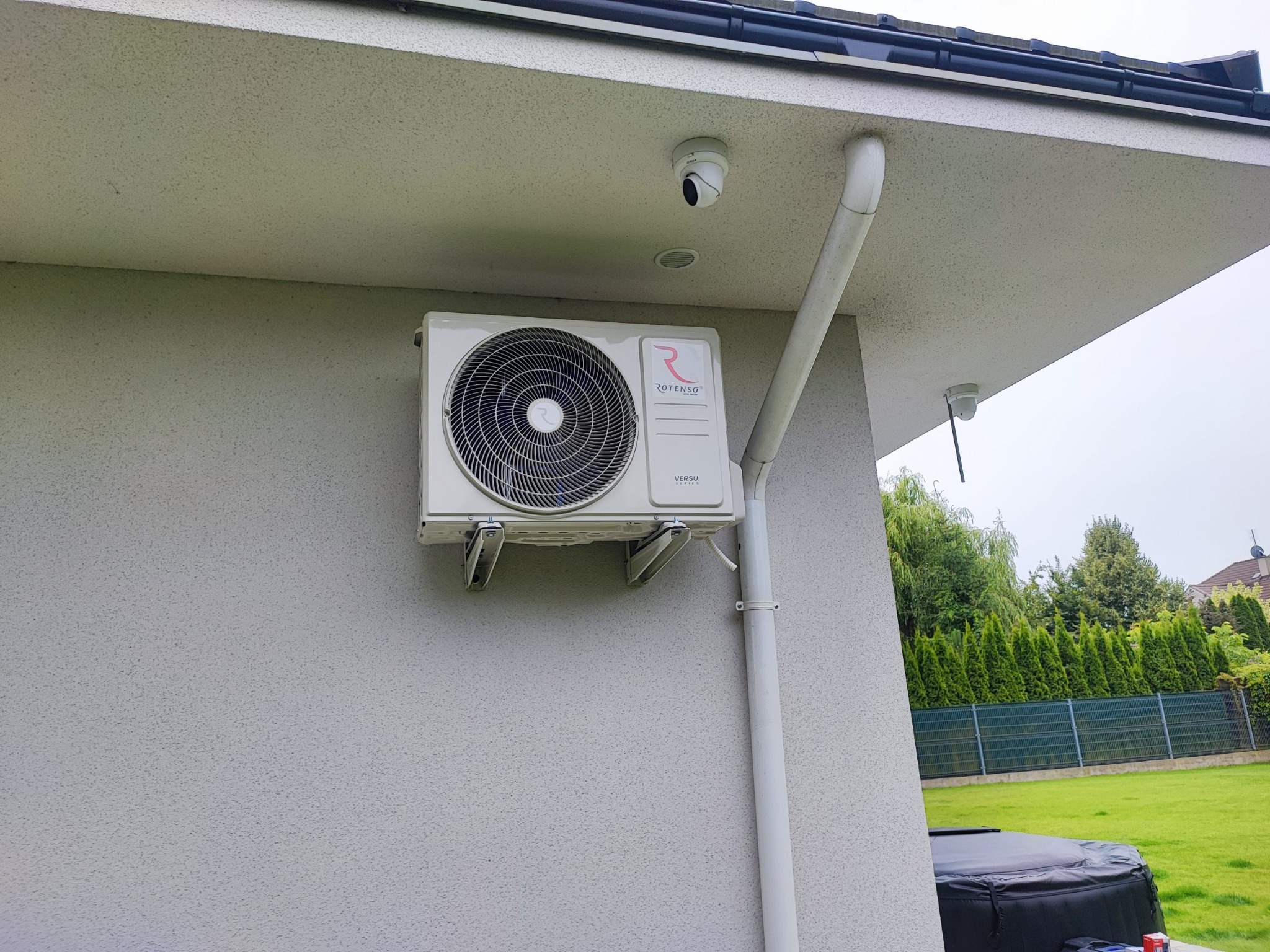 Biała klimatyzacja Rotenso zamontowana na elewacji domu, obok rynna spustowa. Widoczna kamera monitoringu i fragment ogrodu z zielonym trawnikiem.