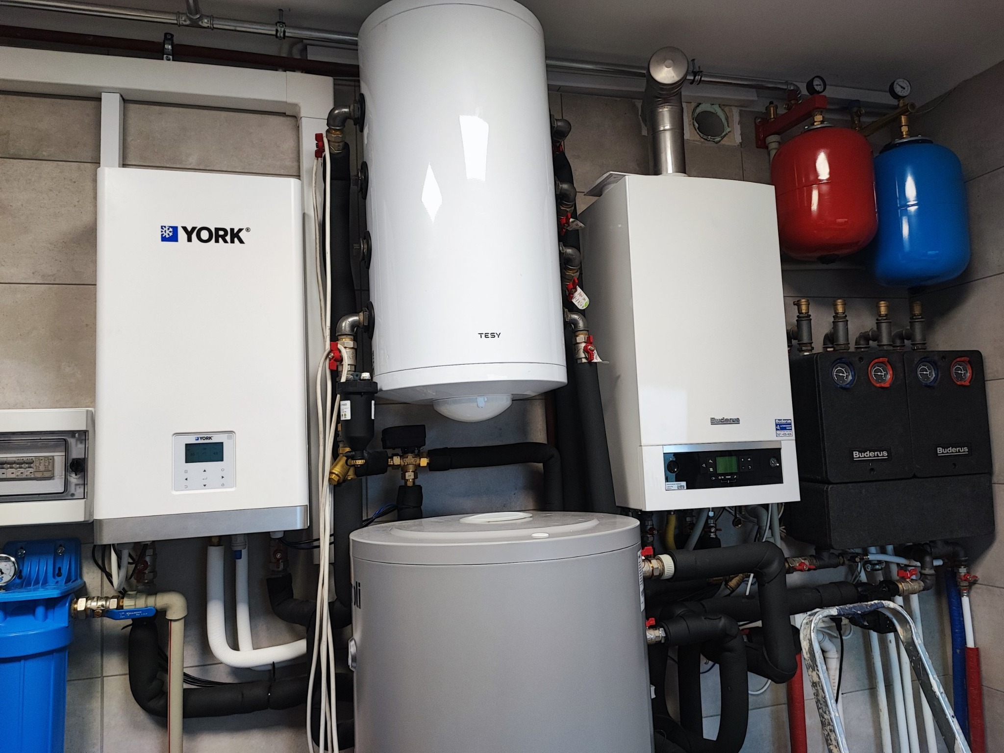 Kompleksowa instalacja grzewcza z kotłami York i Buderus, zasobnikiem Tesy, oraz elementami armatury w pomieszczeniu technicznym z szarą glazurą ścienną.