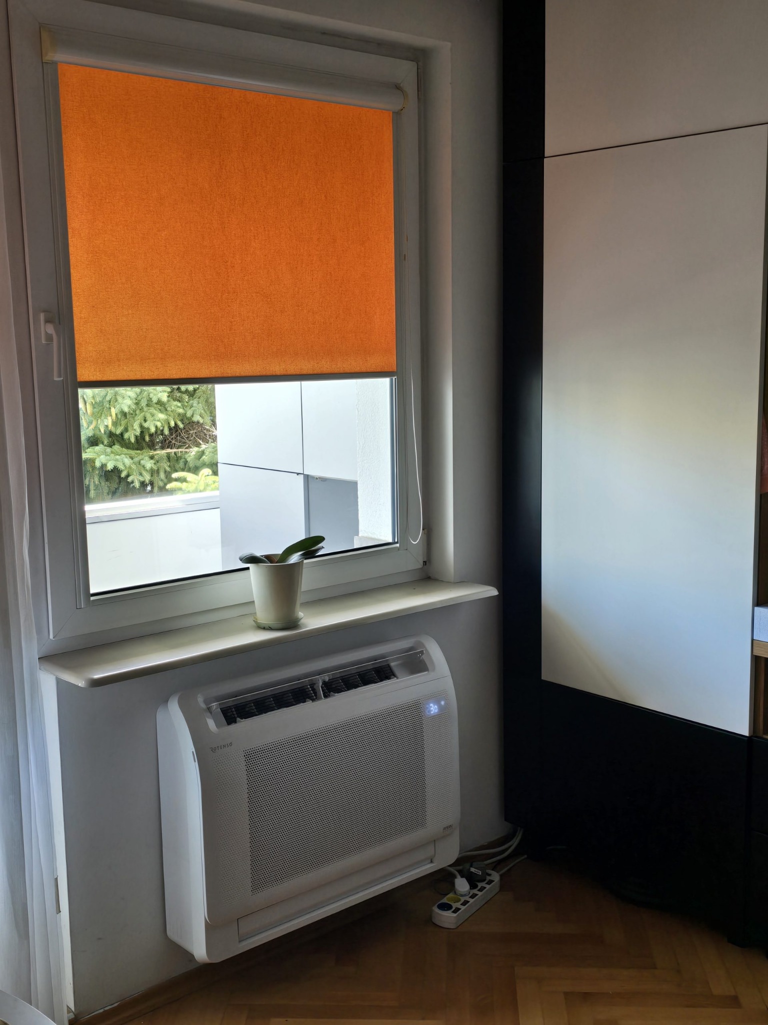 Biały klimatyzator Rotenso zamontowany pod oknem z pomarańczową roletą, obok ciemnego mebla. Widoczny przedłużacz na parkiecie.