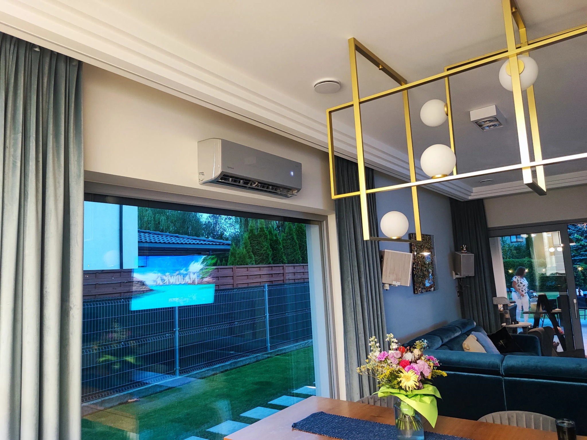 Nowoczesny klimatyzator ścienny w jasnym salonie z widokiem na ogród przez duże okno. Wnętrze dopełnia designerska lampa i eleganckie zasłony.