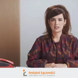 Kobieta w bluzce z geometrycznym wzorem i opasce na włosach, siedząca przy biurku z czerwonym telefonem, prezentuje się na tle jasnej ściany.