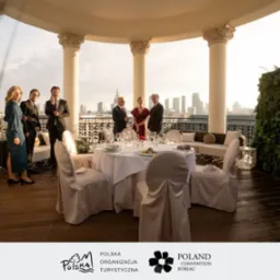 Elegancki event firmowy na tarasie z kolumnami i widokiem na panoramę miasta, uczestnicy w strojach formalnych, stół nakryty do uroczystego obiadu.