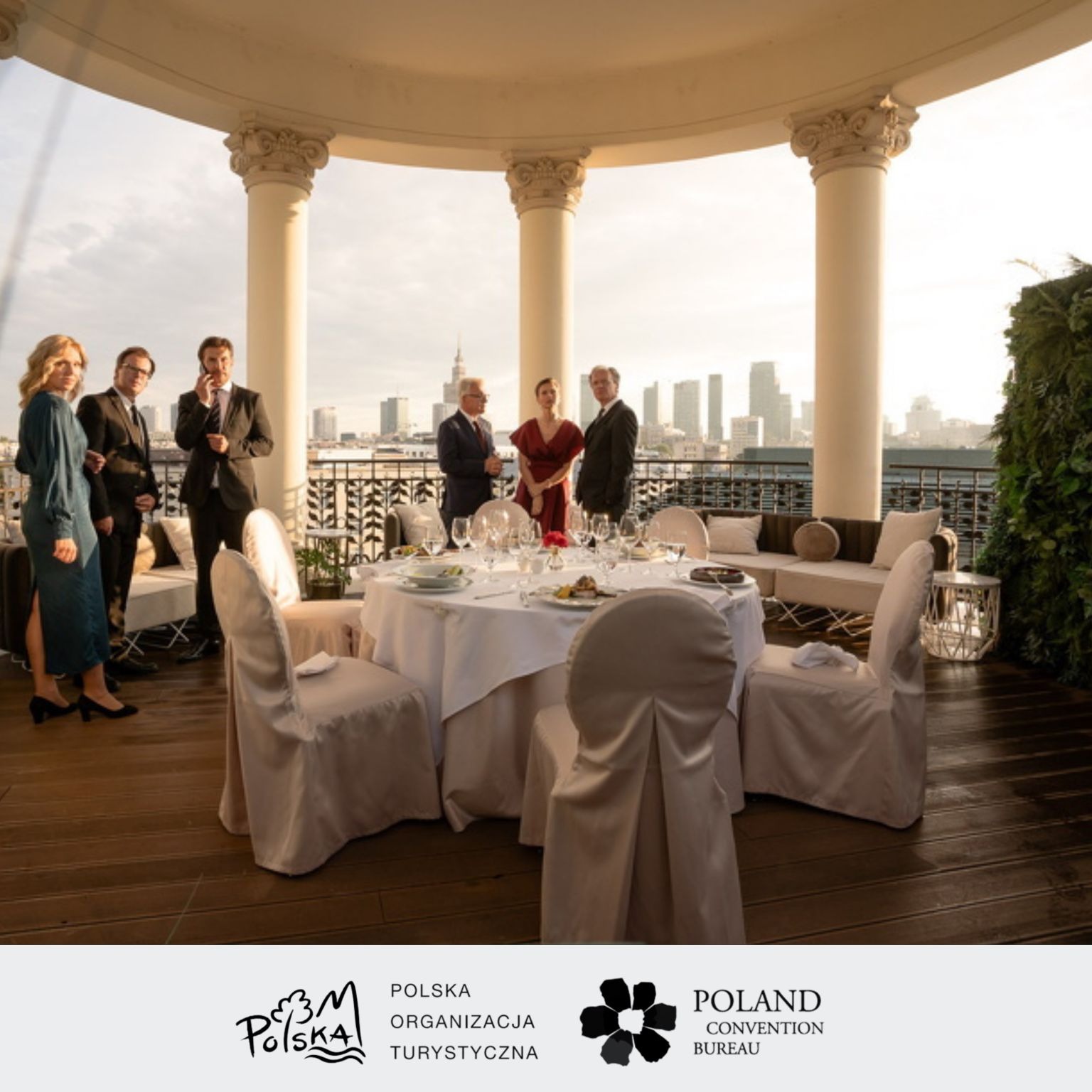 Elegancki event firmowy na tarasie z kolumnami i widokiem na panoramę miasta, uczestnicy w strojach formalnych, stół nakryty do uroczystego obiadu.