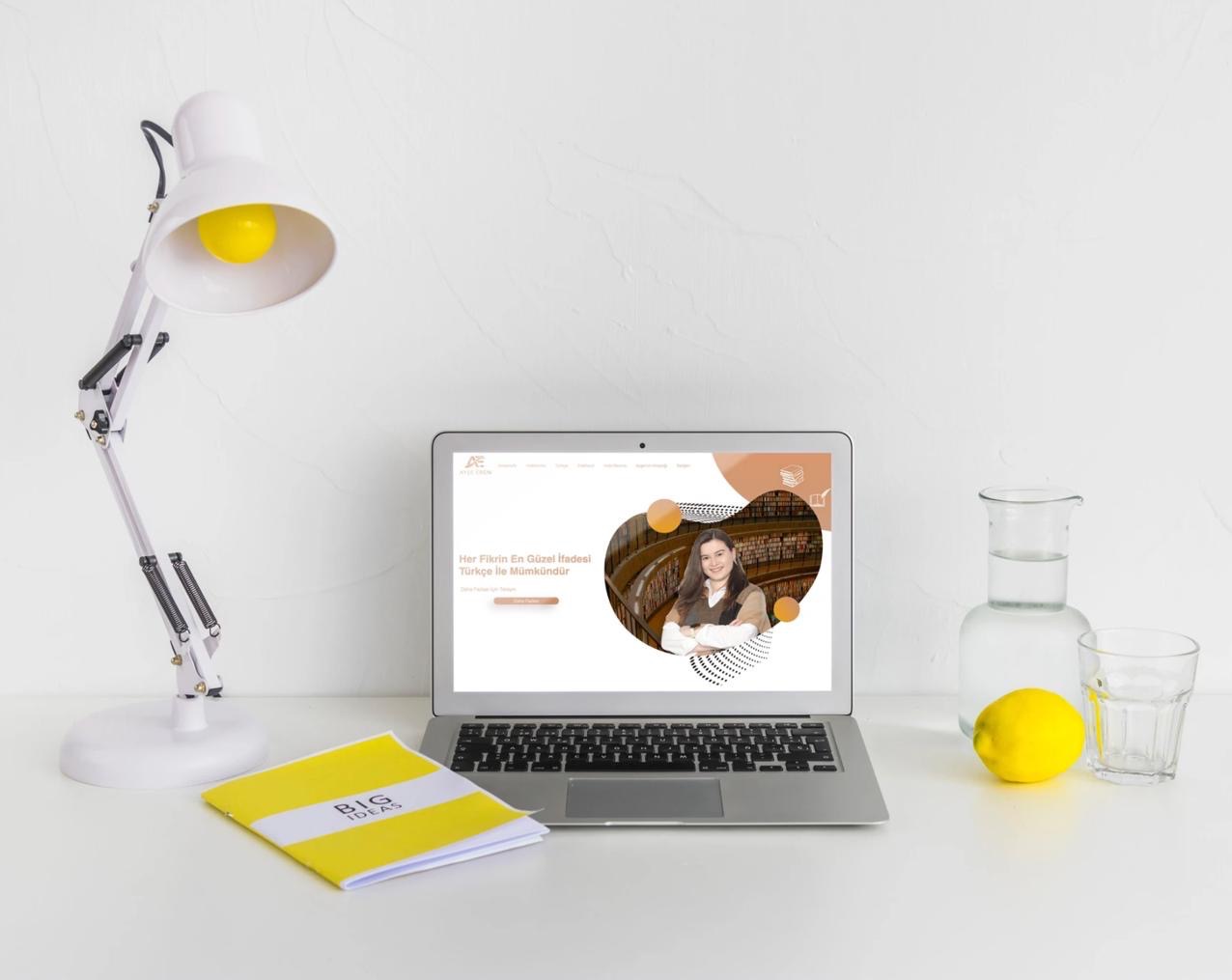Laptop wyświetla stronę internetową z portretem kobiety na tle biblioteki, obok lampka, notatnik 'Big Ideas', dzbanek z wodą i cytryna na białym biurku.