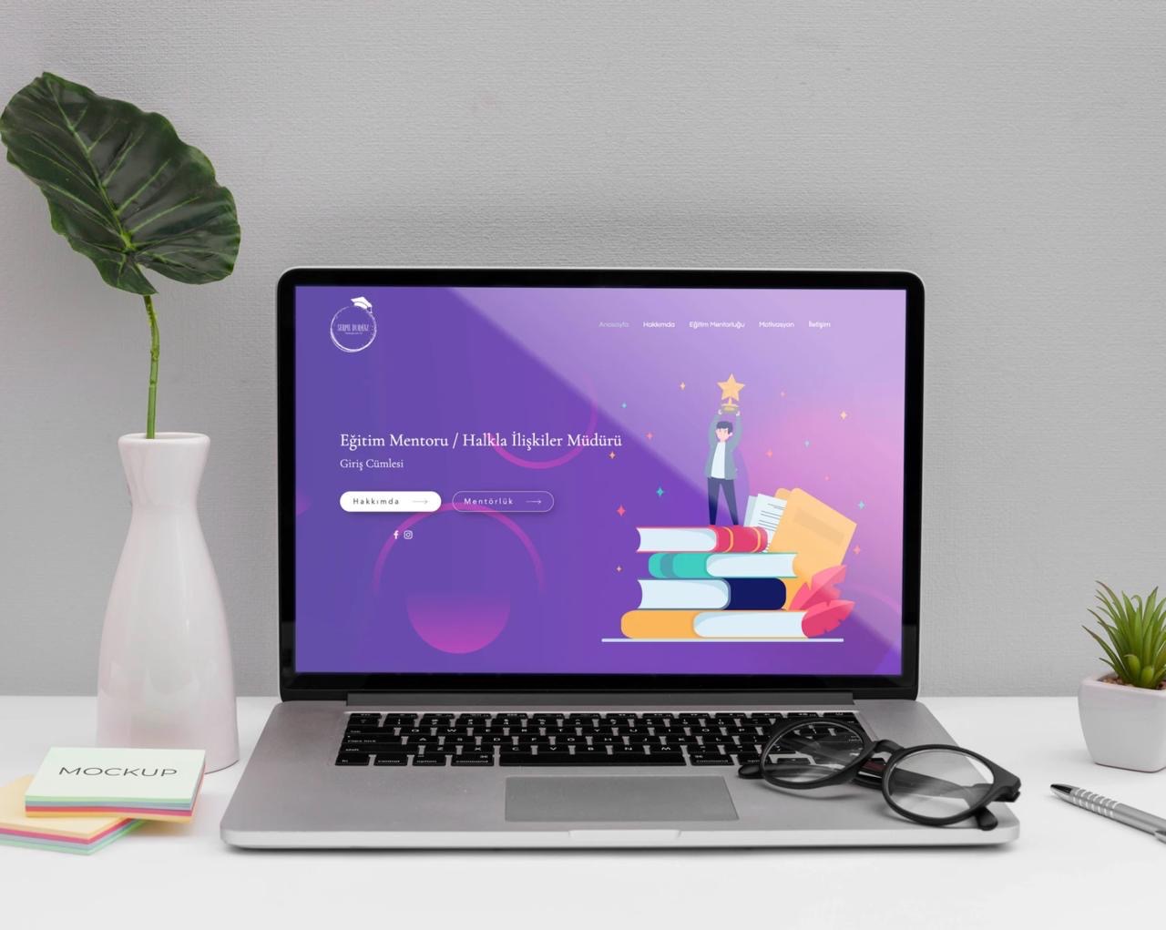 Laptop wyświetlający stronę internetową z ilustracją osoby stojącej na stosie książek, trzymającej gwiazdę, obok wazon z rośliną, notatki samoprzylepne, okulary i długopis na białym biurku.