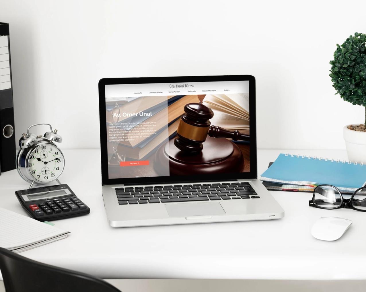Laptop wyświetla stronę internetową kancelarii prawnej z motywem sędziego, obok zegar, kalkulator, notatnik i okulary na białym biurku.