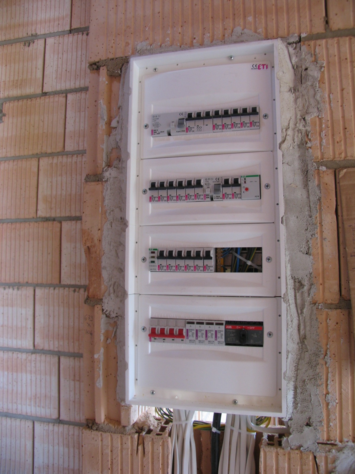 Rozdzielnica elektryczna zamontowana w surowej ścianie z betonu komórkowego, widoczne wystające przewody i niedokończone obrzeża wokół skrzynki