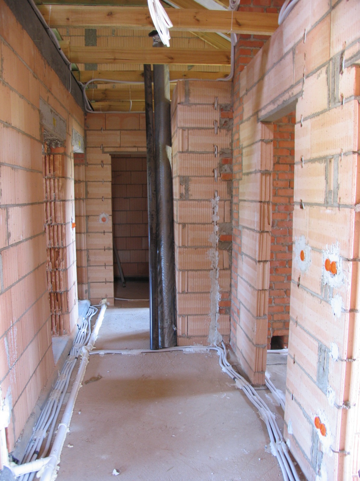Wnętrze w trakcie budowy z widocznymi przewodami elektrycznymi ułożonymi wzdłuż ścian, pomarańczowe puszki instalacyjne w ścianach z cegły, pionowe rury w czarnej folii ochronnej.