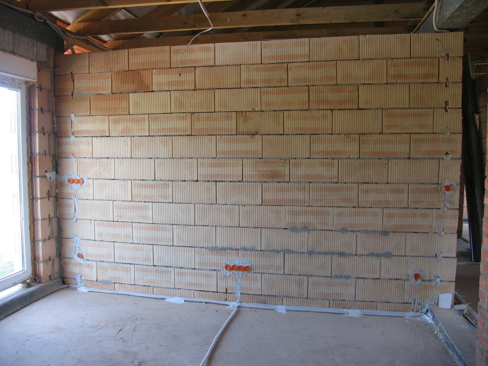 Surowa ściana z pustaków ceramicznych z widocznymi puszkami instalacyjnymi pod gniazdka elektryczne i przewodami w listwach przypodłogowych, w trakcie prac instalacyjnych.