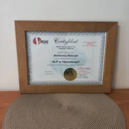 Oprawiony certyfikat ukończenia szkolenia NLP w Hipnoterapii, wystawiony przez Szkołę Hipnozy i Hipnoterapii Sempergo Rafał Foss, z datą 21-04-2024, leżący na okrągłej podkładce.