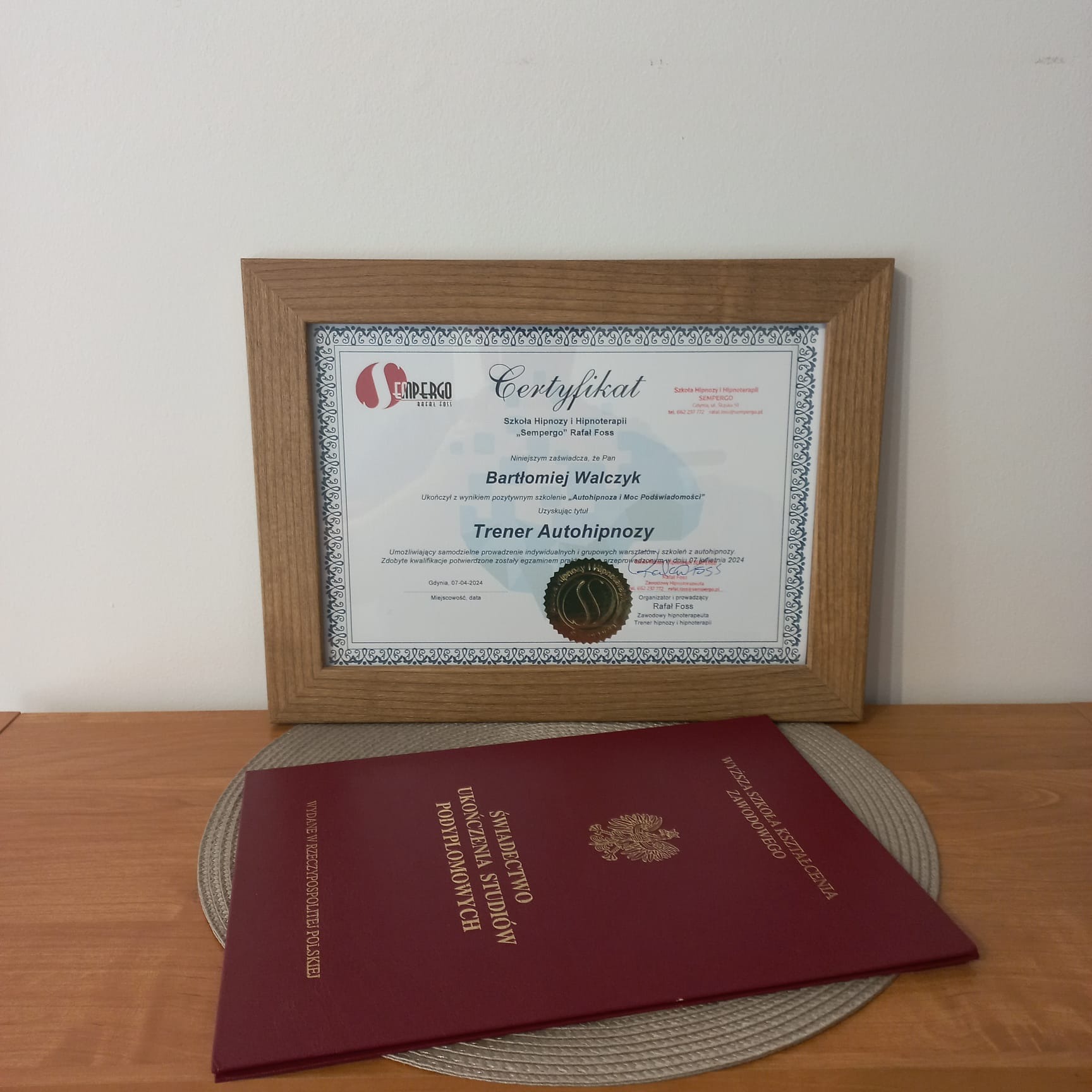 Certyfikat ukończenia kursu 'Trener Autohipnozy' w ramce, obok świadectwo ukończenia studiów podyplomowych na drewnianym stole.