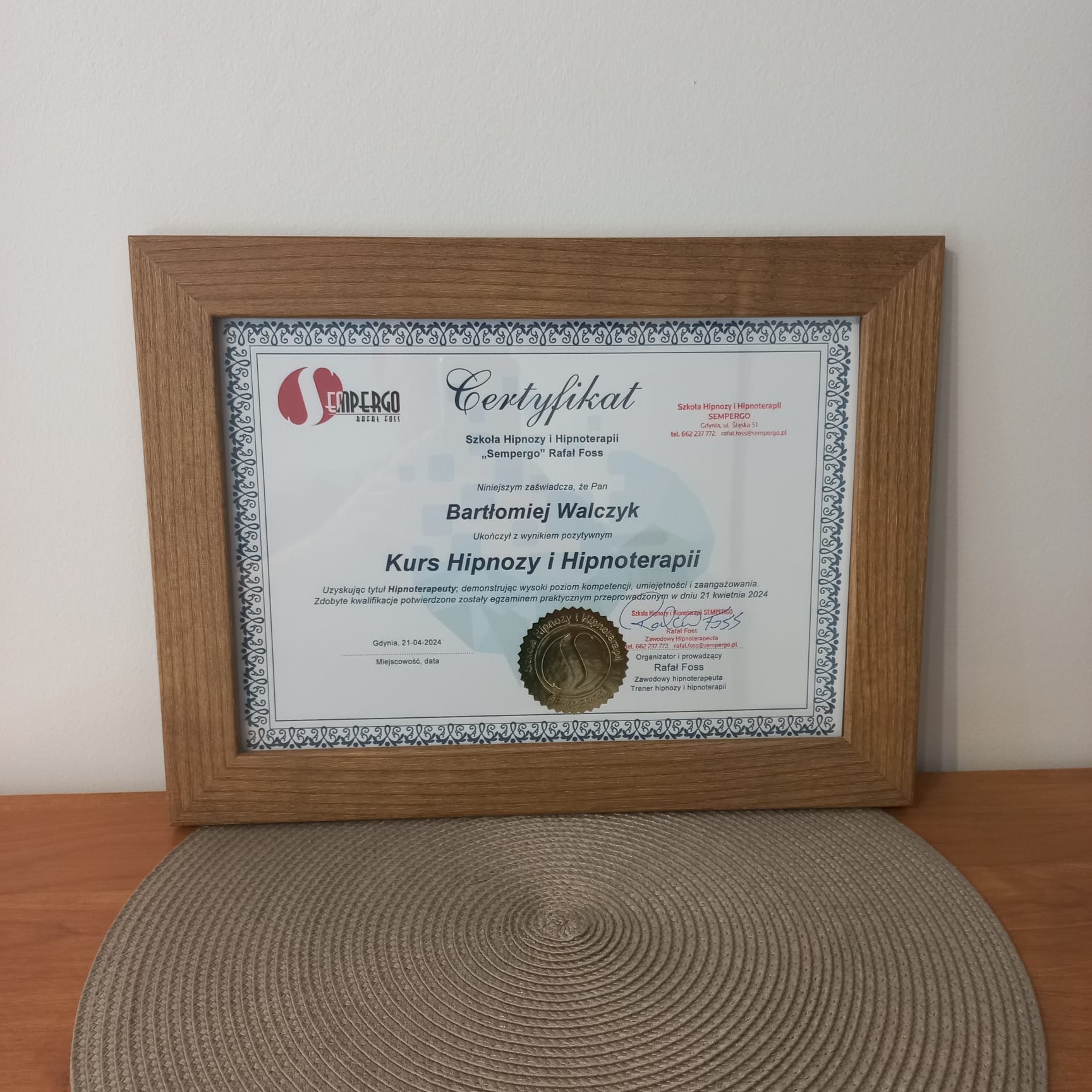 Oprawiony w drewnianą ramkę certyfikat ukończenia kursu hipnozy i hipnoterapii Szkoły Hipnozy i Hipnoterapii 'Sempergo' Rafała Fossa, leżący na okrągłej, plecionej podkładce.