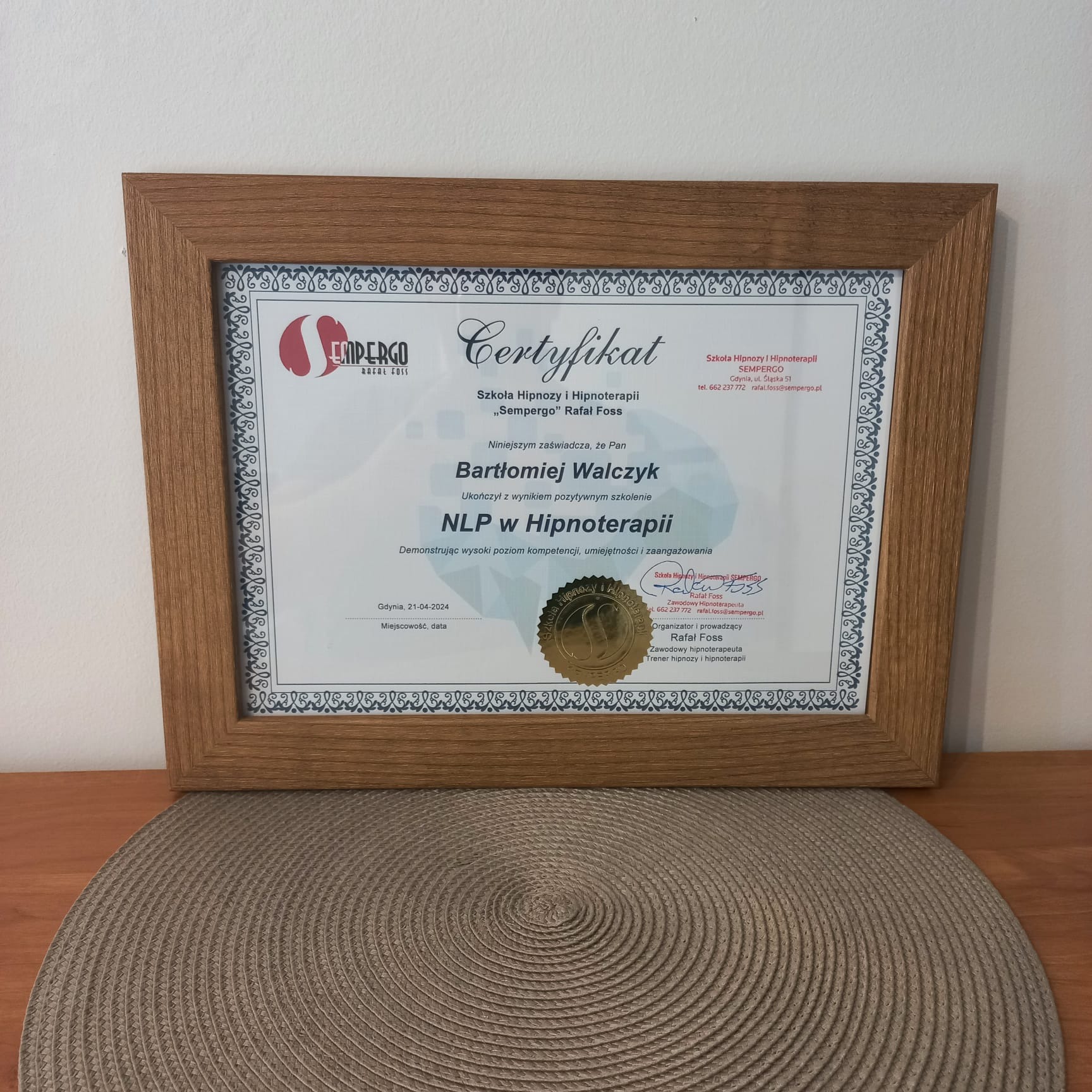 Oprawiony certyfikat ukończenia szkolenia NLP w Hipnoterapii, wystawiony przez Szkołę Hipnozy i Hipnoterapii Sempergo Rafał Foss, z datą 21-04-2024, leżący na okrągłej podkładce.