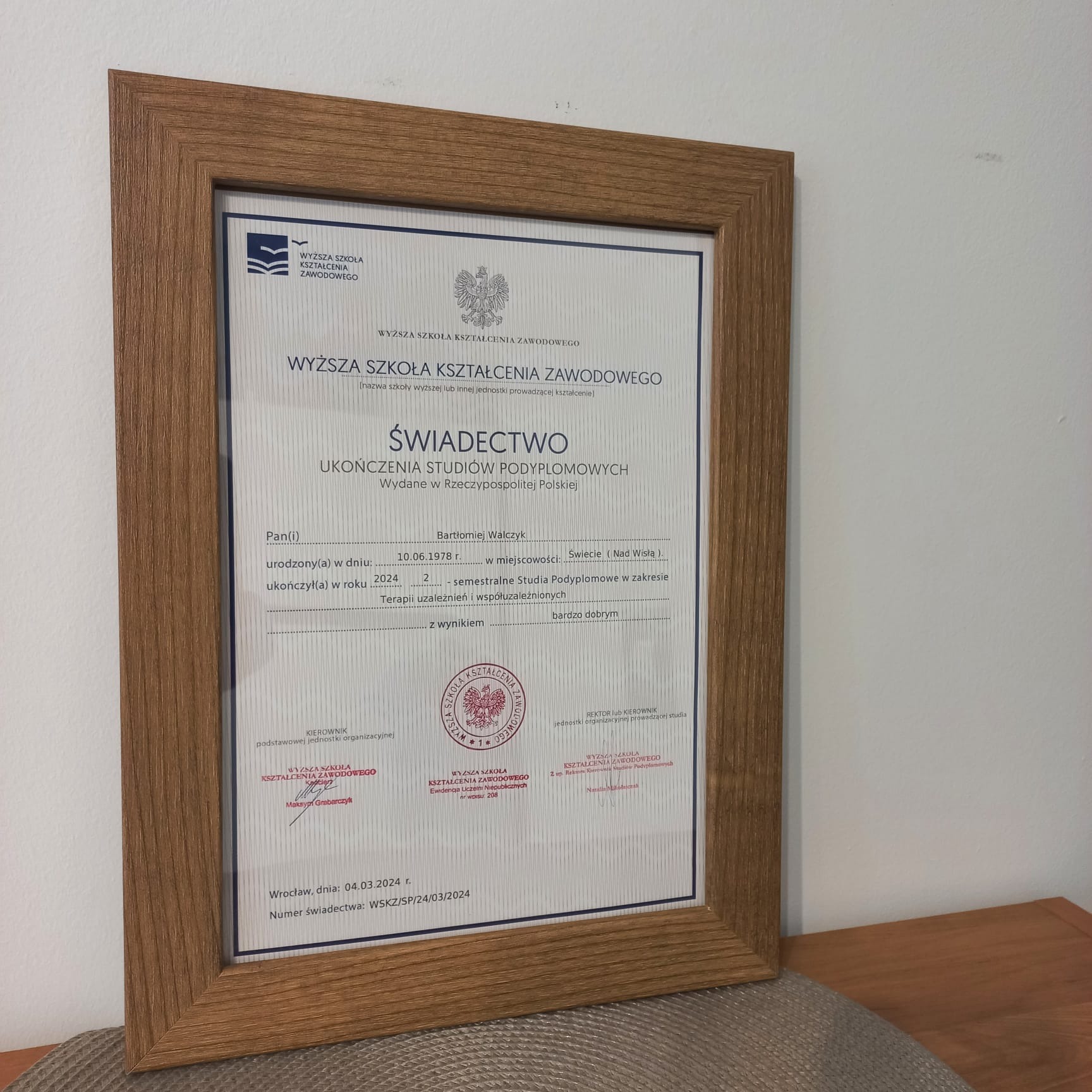 Oprawione w drewnianą ramę świadectwo ukończenia studiów podyplomowych w zakresie terapii uzależnień, wydane przez Wyższą Szkołę Kształcenia Zawodowego, z datą 04.03.2024.
