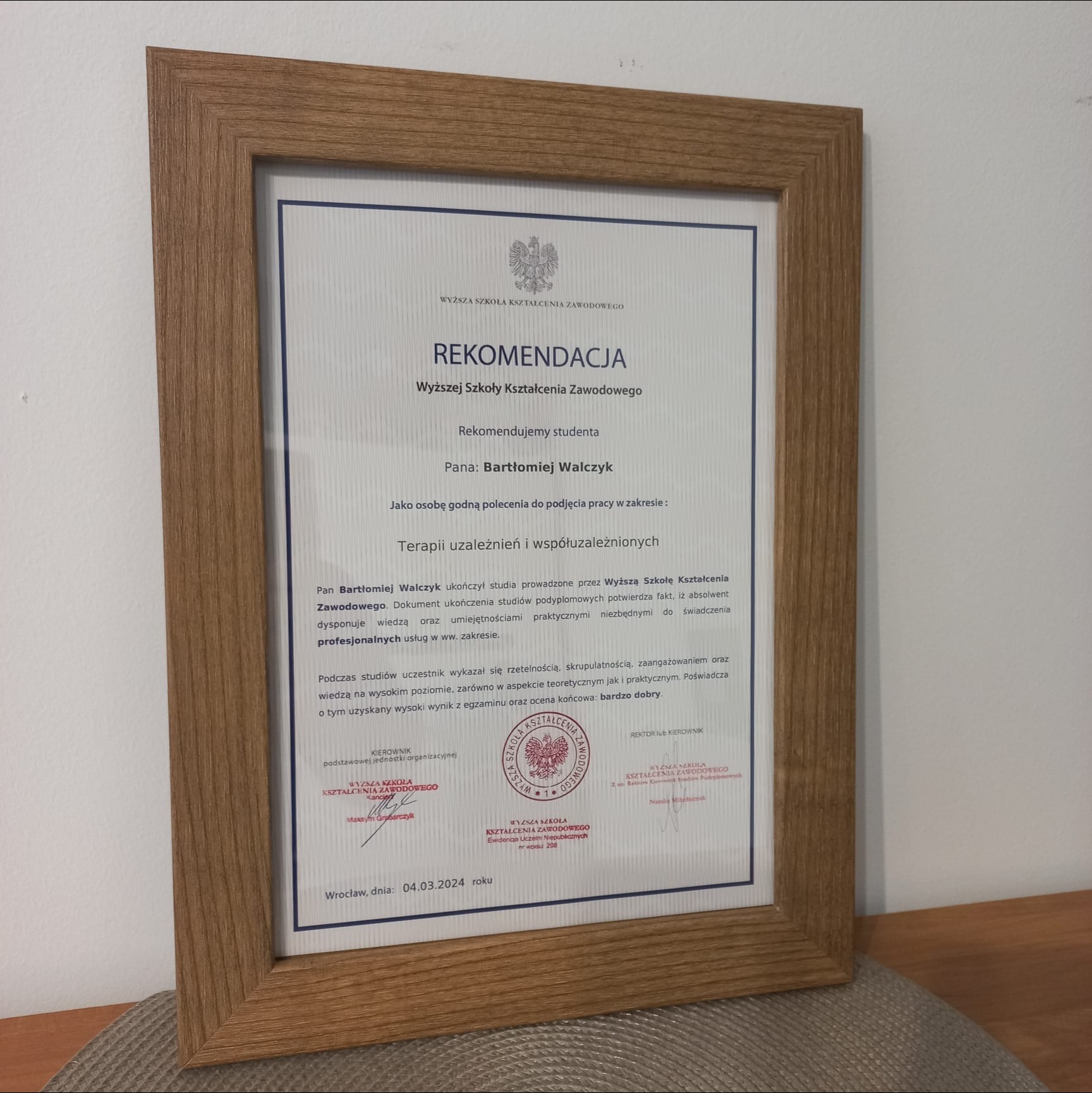 Oprawiona w drewnianą ramkę rekomendacja Wyższej Szkoły Kształcenia Zawodowego dla Bartłomieja Walczyka w zakresie terapii uzależnień, datowana na 04.03.2024, umieszczona na stole.