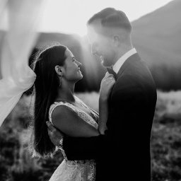 Zebra fotografia - Czarno-białe zdjęcie nowożeńców w objęciach na tle gór, z welonem powiewającym na wietrze i promieniami słońca za nimi.