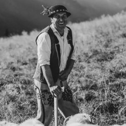 Zebra fotografia - Czarno-białe zdjęcie uśmiechniętego mężczyzny w tradycyjnym góralskim stroju, z kapeluszem ozdobionym piórami, trzymającego laskę, na tle górskiego krajobrazu i stada owiec.