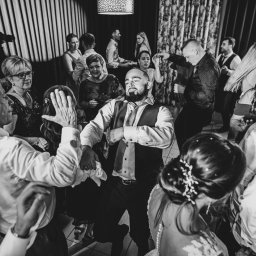 Zebra fotografia - Czarno-białe zdjęcie z wesela: goście tańczą na parkiecie, w tle fotograf z dużym obiektywem, mężczyzna w środku kadru w koszuli i kamizelce z krawatem, zamyślony wyraz twarzy.