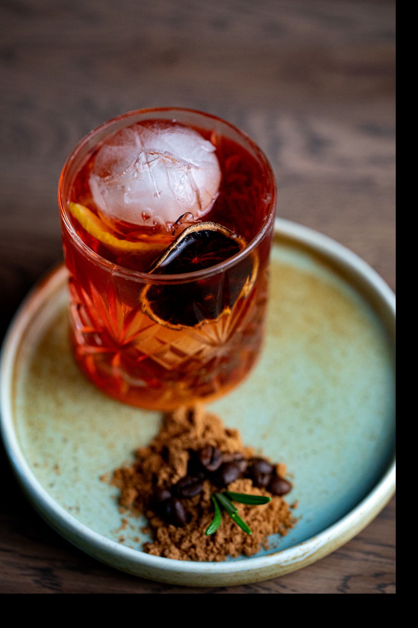 Kryształowy kieliszek z czerwonym koktajlem, dużą kulą lodu, suszoną pomarańczą i skórką cytryny, podany na talerzu z kawą mieloną, ziarnami kawy i gałązką rozmarynu.