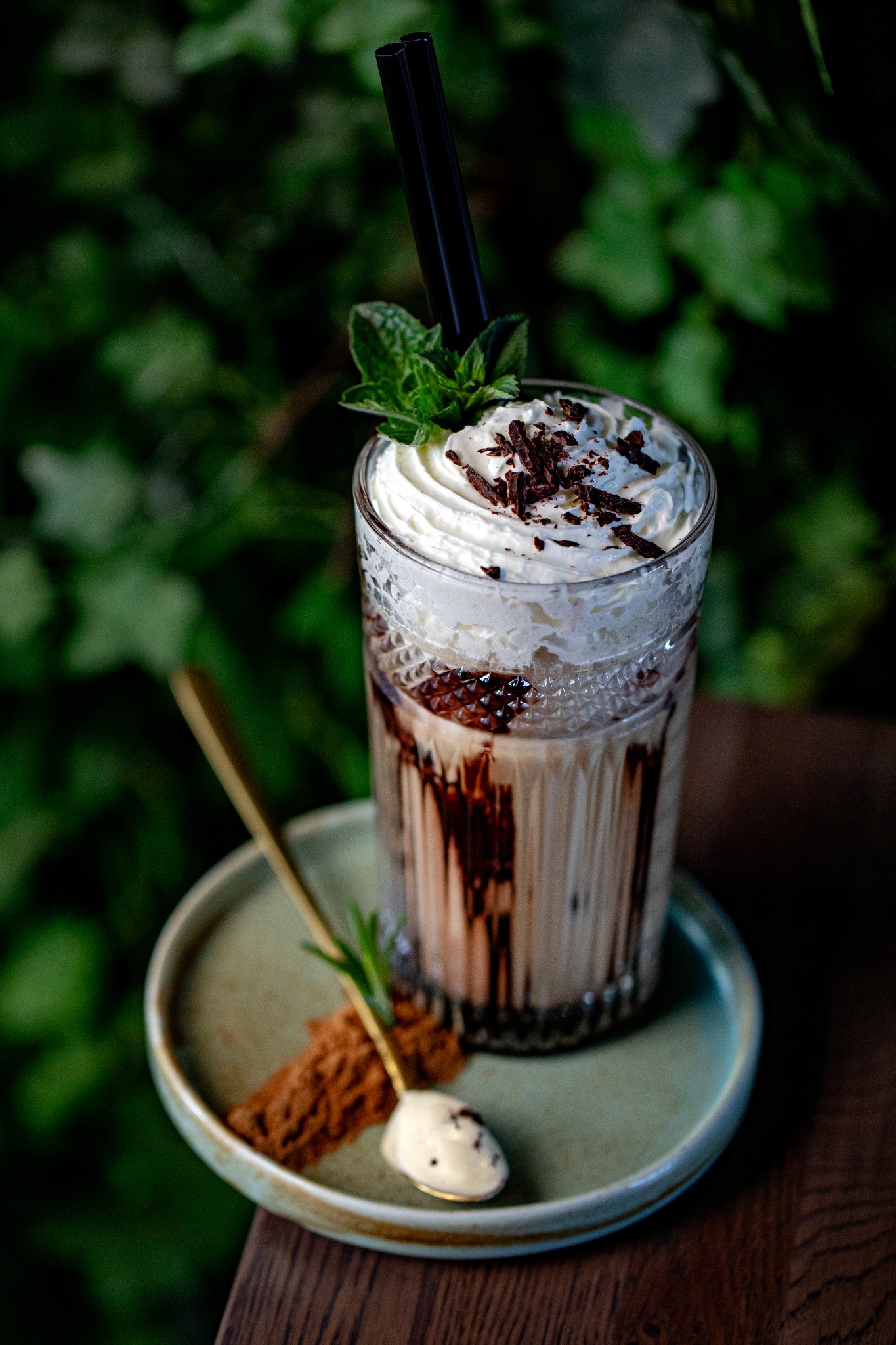 Dekoracyjny szklany kieliszek z kawą mrożoną, bitą śmietaną, posypką czekoladową i miętą, umieszczony na małym talerzyku z ciasteczkiem i złotą łyżeczką, na tle zieleni.