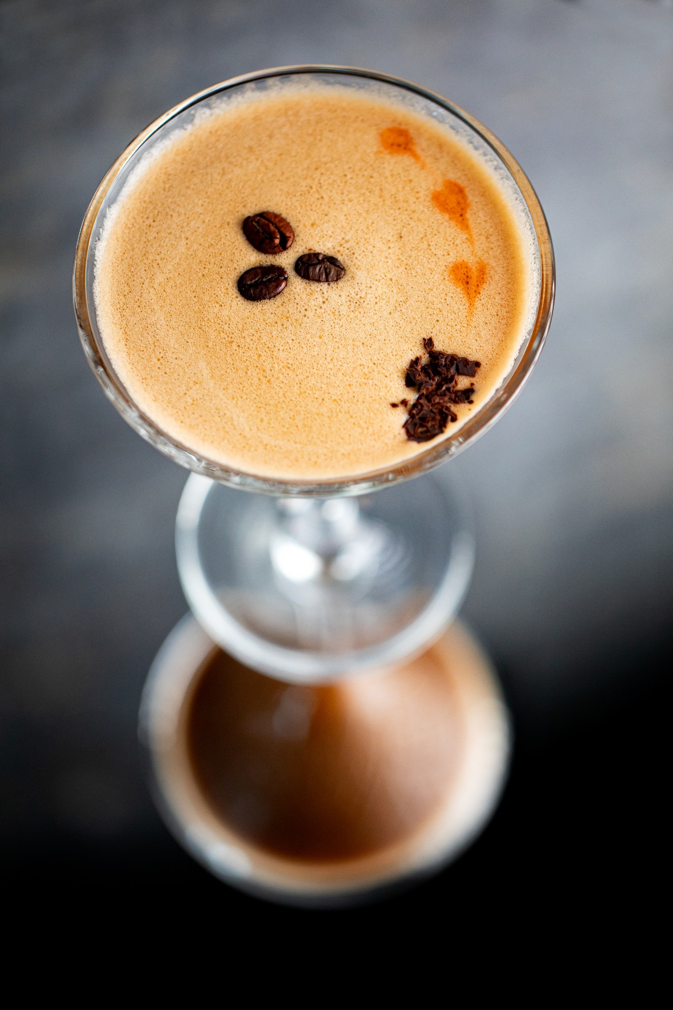 Espresso martini w kieliszku koktajlowym z dekoracją z trzech ziaren kawy i posypką z ciemnej czekolady, widok z góry na ciemnym tle.