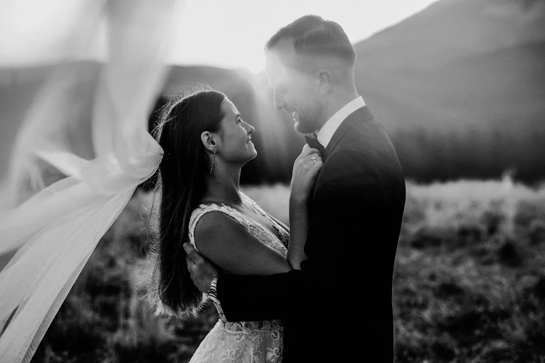 Czarno-białe zdjęcie nowożeńców w objęciach na tle gór, z welonem powiewającym na wietrze i promieniami słońca za nimi.