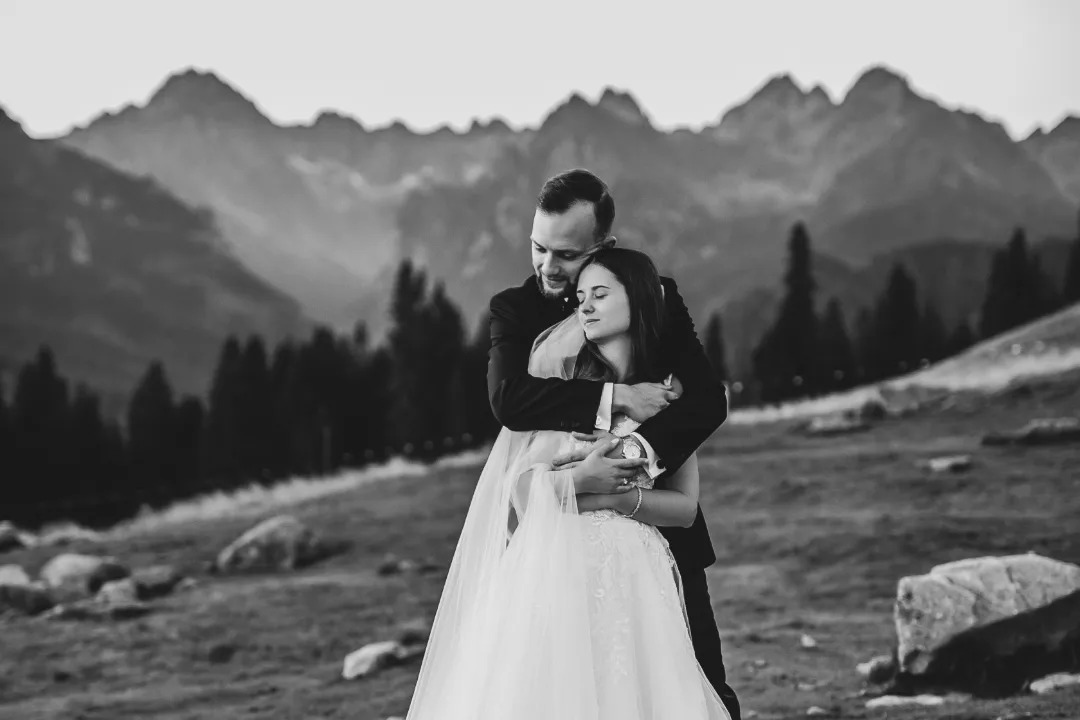 Czarno-białe zdjęcie nowożeńców w objęciach na tle górskiego krajobrazu z lasem iglastym i mglistymi szczytami.
