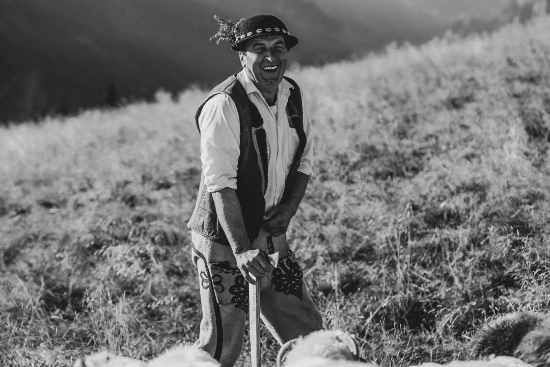 Czarno-białe zdjęcie uśmiechniętego mężczyzny w tradycyjnym góralskim stroju, z kapeluszem ozdobionym piórami, trzymającego laskę, na tle górskiego krajobrazu i stada owiec.
