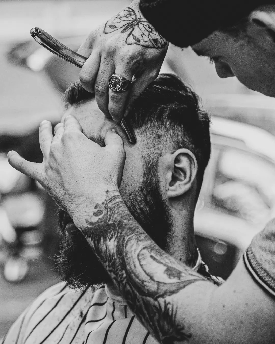 Czarno-białe zdjęcie przedstawia fryzjera golącego brzytwą czoło klienta z brodą, widoczne tatuaże na rękach obu mężczyzn, pierścień z drzewem na palcu fryzjera.