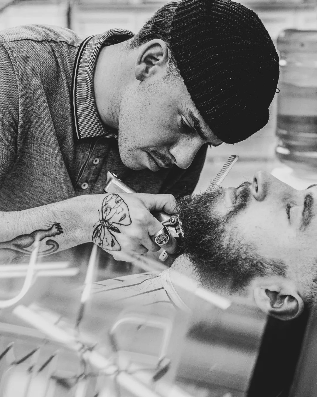 Czarno-białe zdjęcie fryzjera w czapce, który z bliska strzyże brodę klienta leżącego na fotelu, widoczne tatuaże na ręce fryzjera.