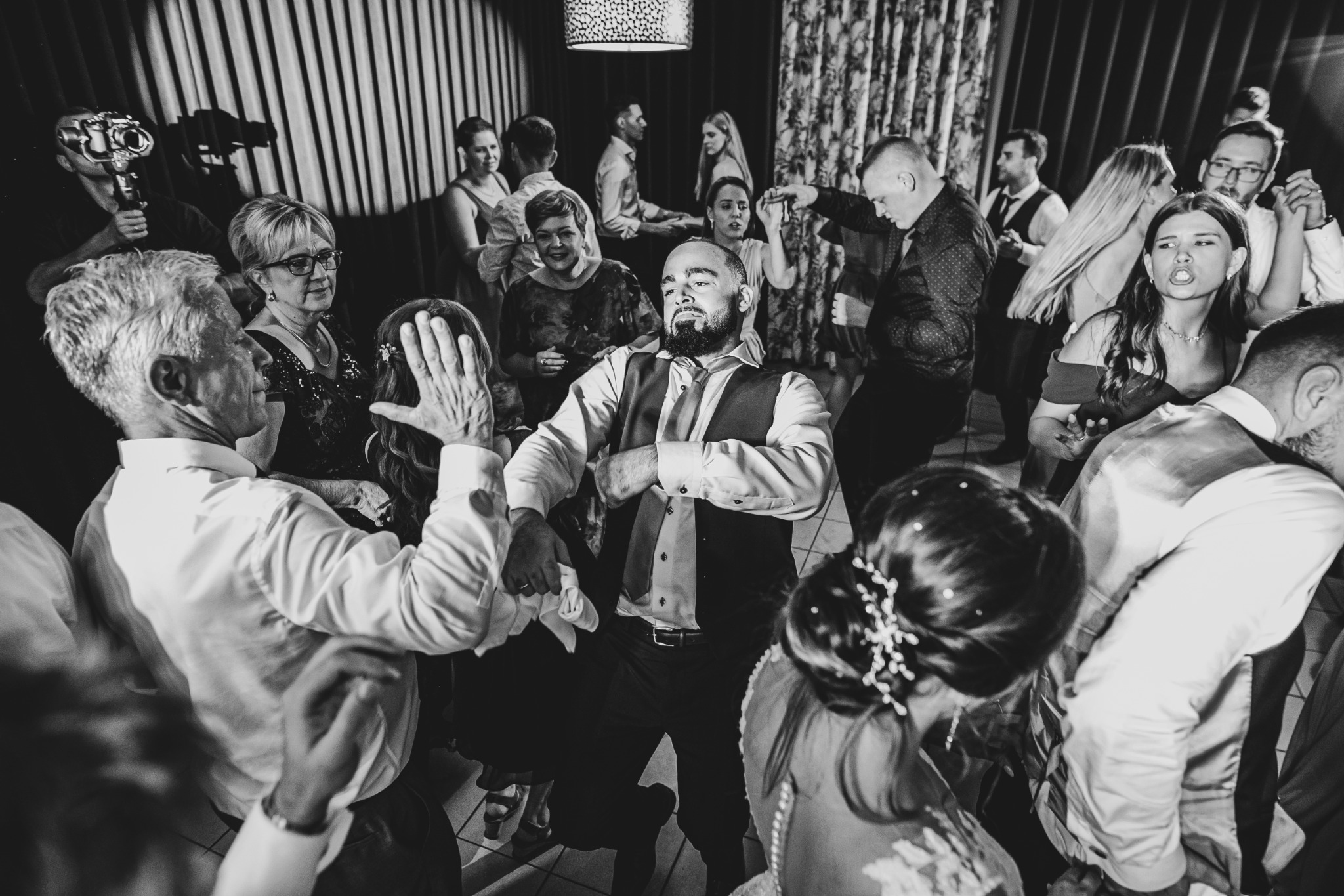 Czarno-białe zdjęcie z wesela: goście tańczą na parkiecie, w tle fotograf z dużym obiektywem, mężczyzna w środku kadru w koszuli i kamizelce z krawatem, zamyślony wyraz twarzy.