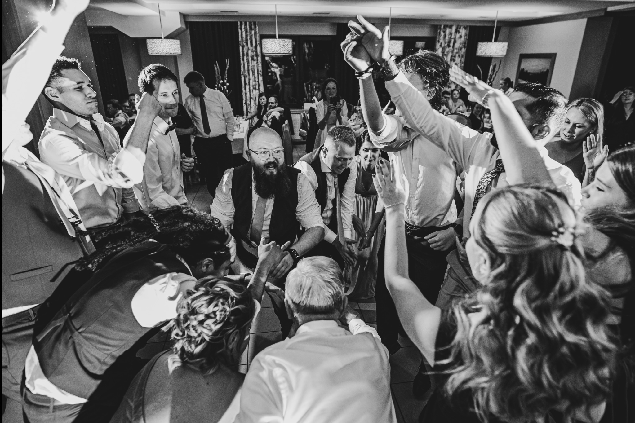Czarno-białe zdjęcie z góry przedstawiające grupę gości weselnych otaczających parę młodą podczas oczepin w sali bankietowej.